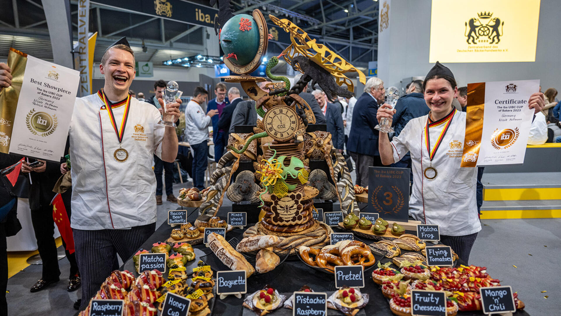 Zwei Köche feiern mit Medaillen, Trophäen und Urkunden vor einer kunstvollen Brot- und Gebäckauslage mit einer Weltkugel, einem Dinosaurier und einer Uhr, umgeben von etikettierten Backwaren bei einer Wettbewerbsveranstaltung.