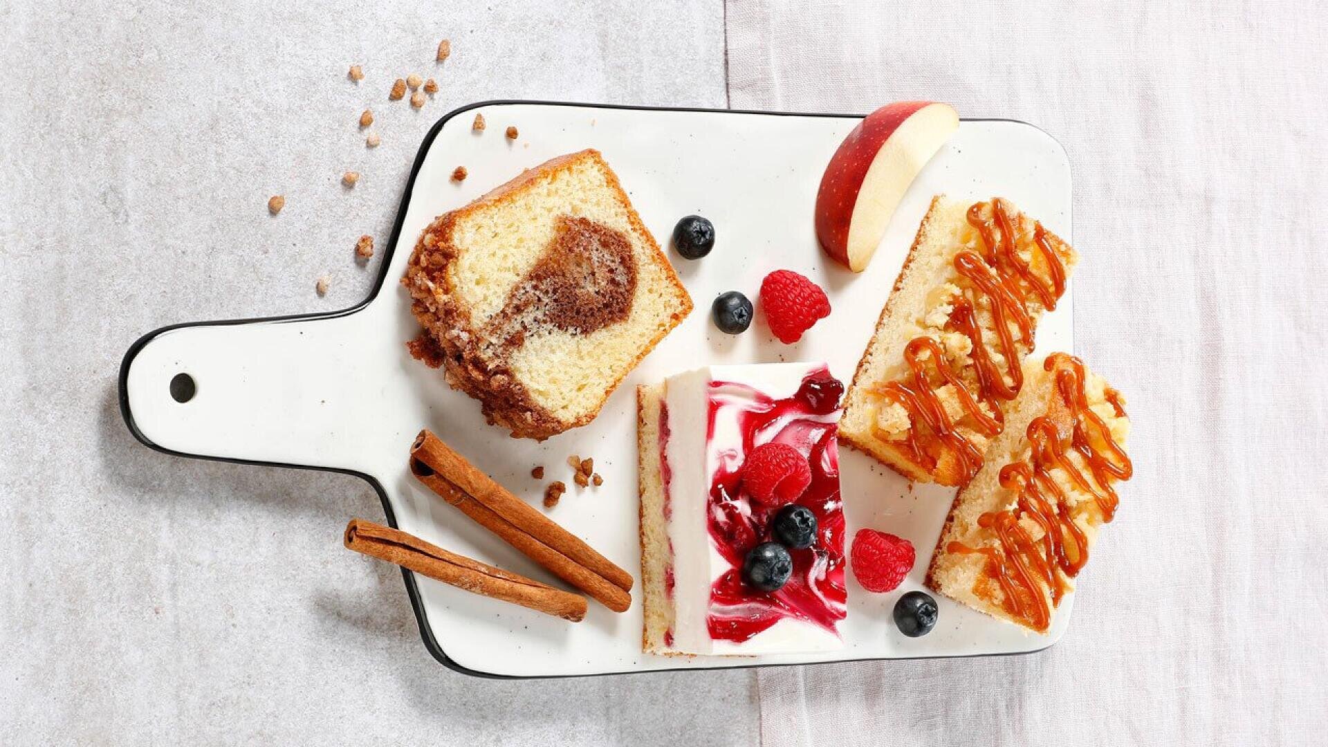A white serving board holds slices of marble cake, caramel-topped cake, berry cheesecake, fresh raspberries, blueberries, an apple wedge, and two cinnamon sticks on a light background.