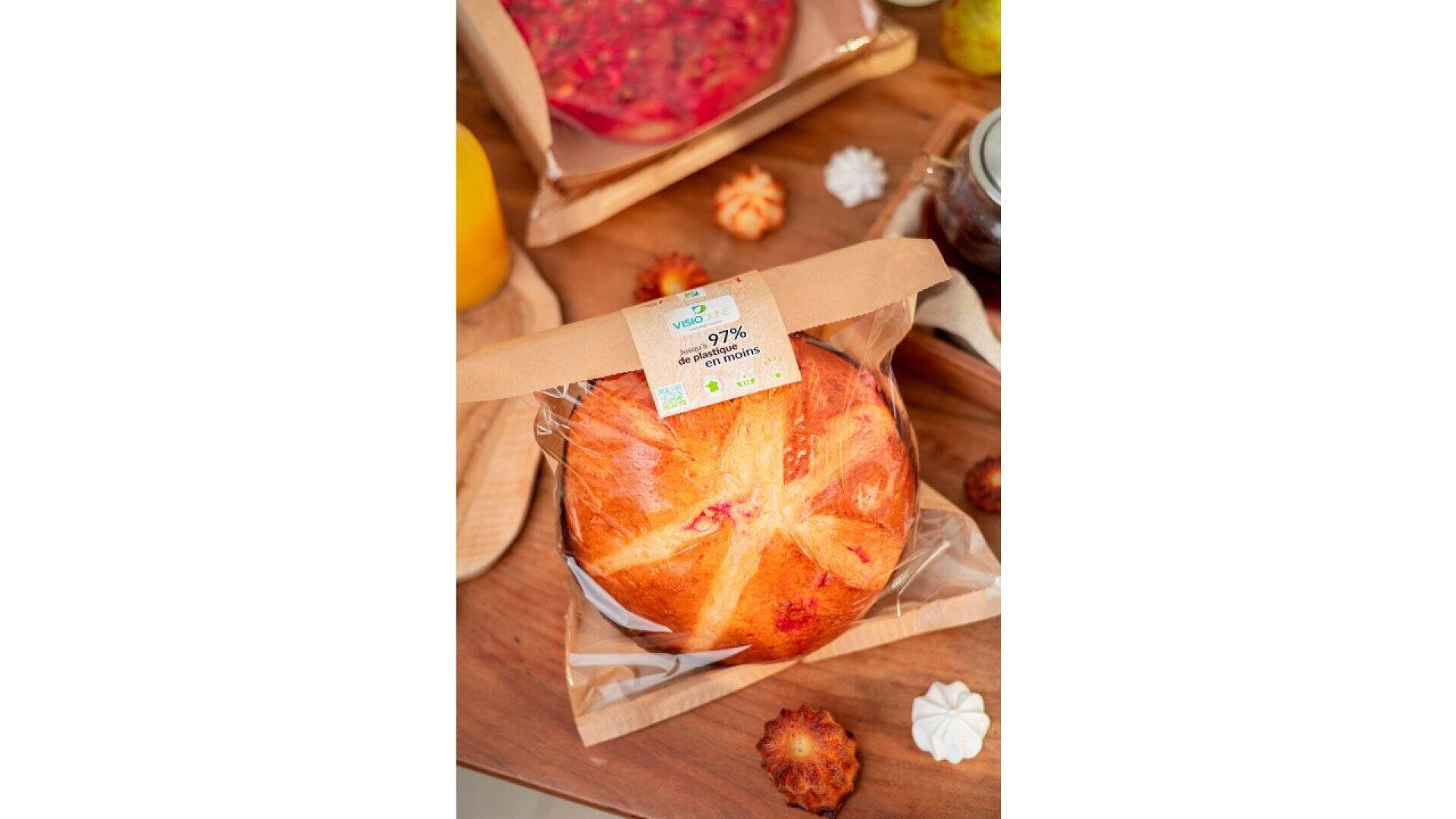 Ein runder Brotlaib in einer durchsichtigen Verpackung mit einem braunen Papieretikett liegt auf einem Holztisch neben Gebäck, einem Getränk und einem Einmachglas. Das Brot hat eine goldene Kruste und ist mit einem Kreuz auf der Oberseite gekennzeichnet.