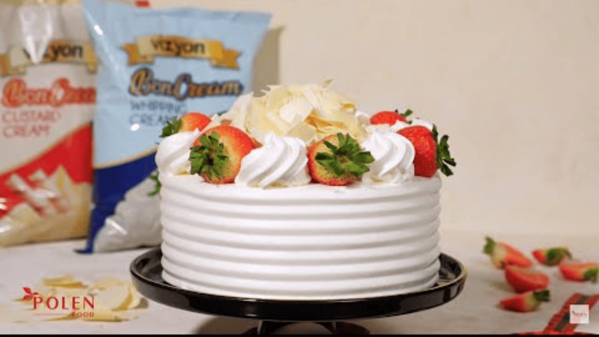 Ein runder weißer Gugelhupf mit Schlagsahne, frischen Erdbeeren und weißen Schokoladenkringeln steht auf einem schwarzen Kuchenständer. Im Hintergrund sind geschnittene Erdbeeren und Dessertmischungen zu sehen.