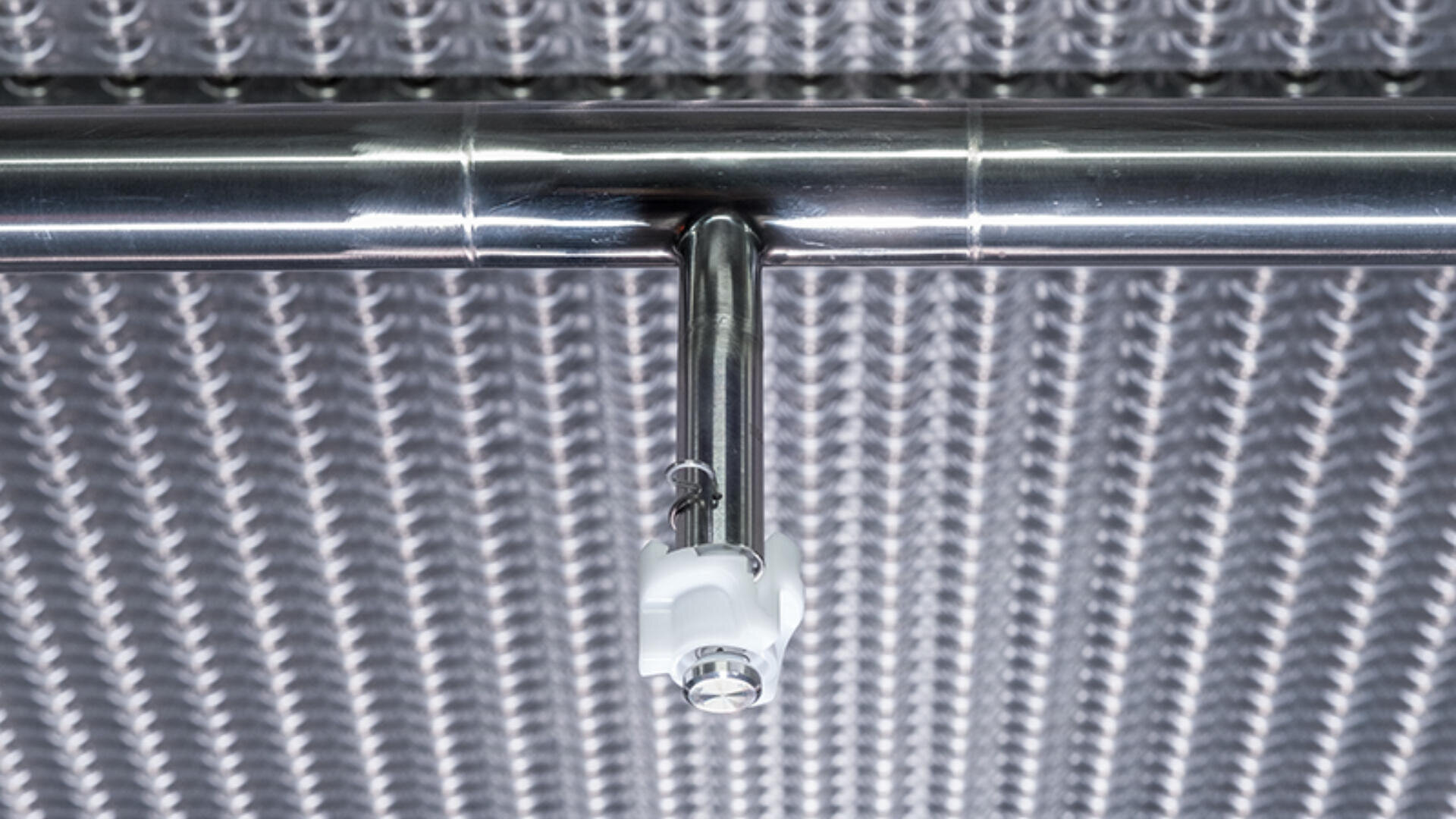 Close-up of a stainless steel sprinkler head attached to a metal pipe, mounted on a ceiling with a textured, metallic surface in the background.