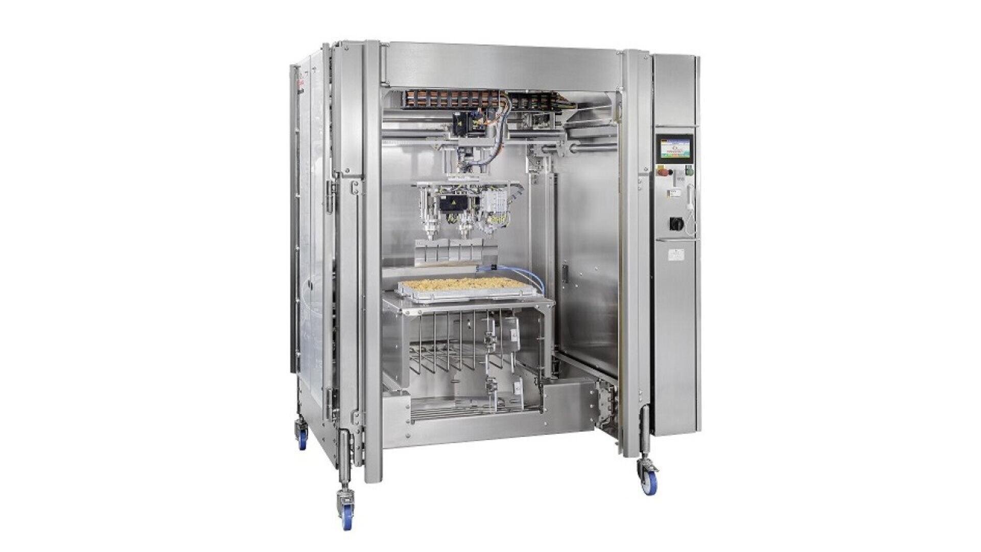 A large industrial stainless steel food processing machine with an open front, showing automated components and a tray holding food inside. The machine stands on four wheels and has a control panel on the right side.