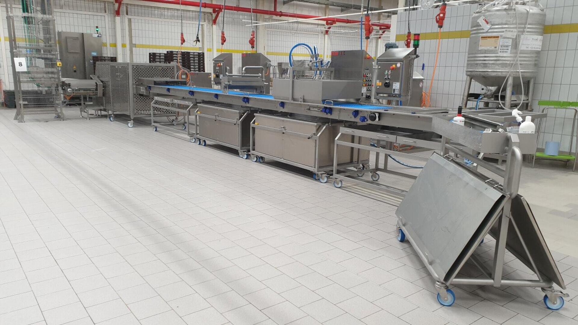 A modern industrial food processing line with stainless steel equipment and conveyor belts in a clean, tiled factory setting. Various machines are connected, and containers and tools are nearby.
