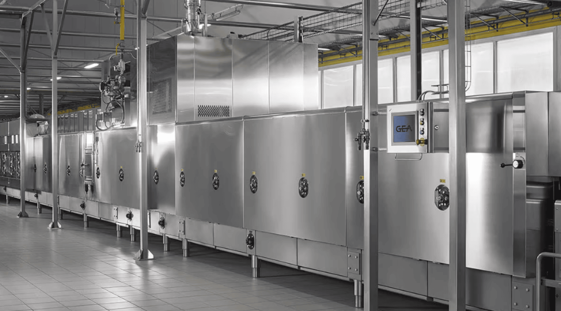 Eine große industrielle Lebensmittelverarbeitungsmaschine aus Edelstahl mit Schalttafeln und Rohrleitungen in einer sauberen, gekachelten Fabrikumgebung.