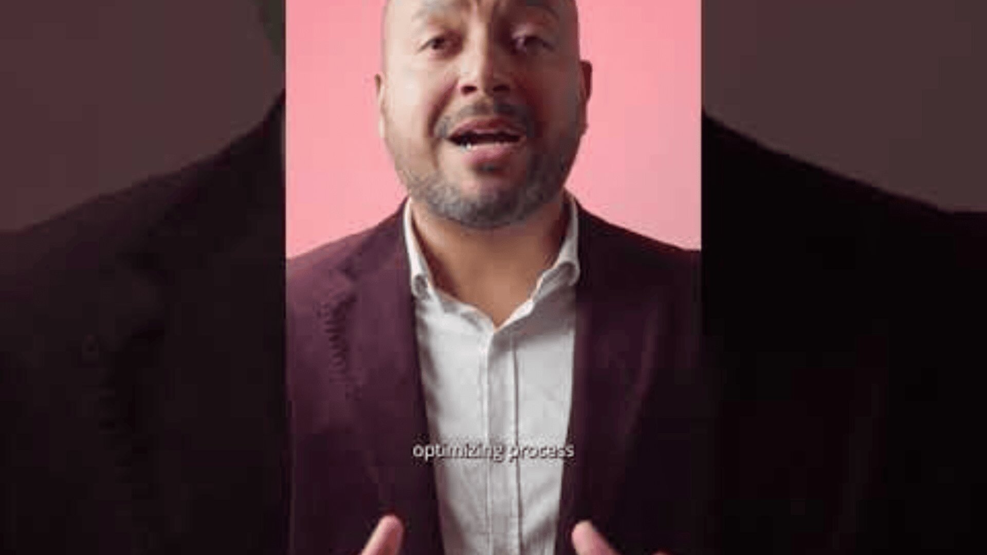 Ein Mann mit gestutztem Bart, einem kastanienbraunen Blazer und einem hellen Hemd spricht und gestikuliert dabei mit den Händen vor einem rosa Hintergrund. Die Untertitel unten lauten „Optimierungsprozess“.