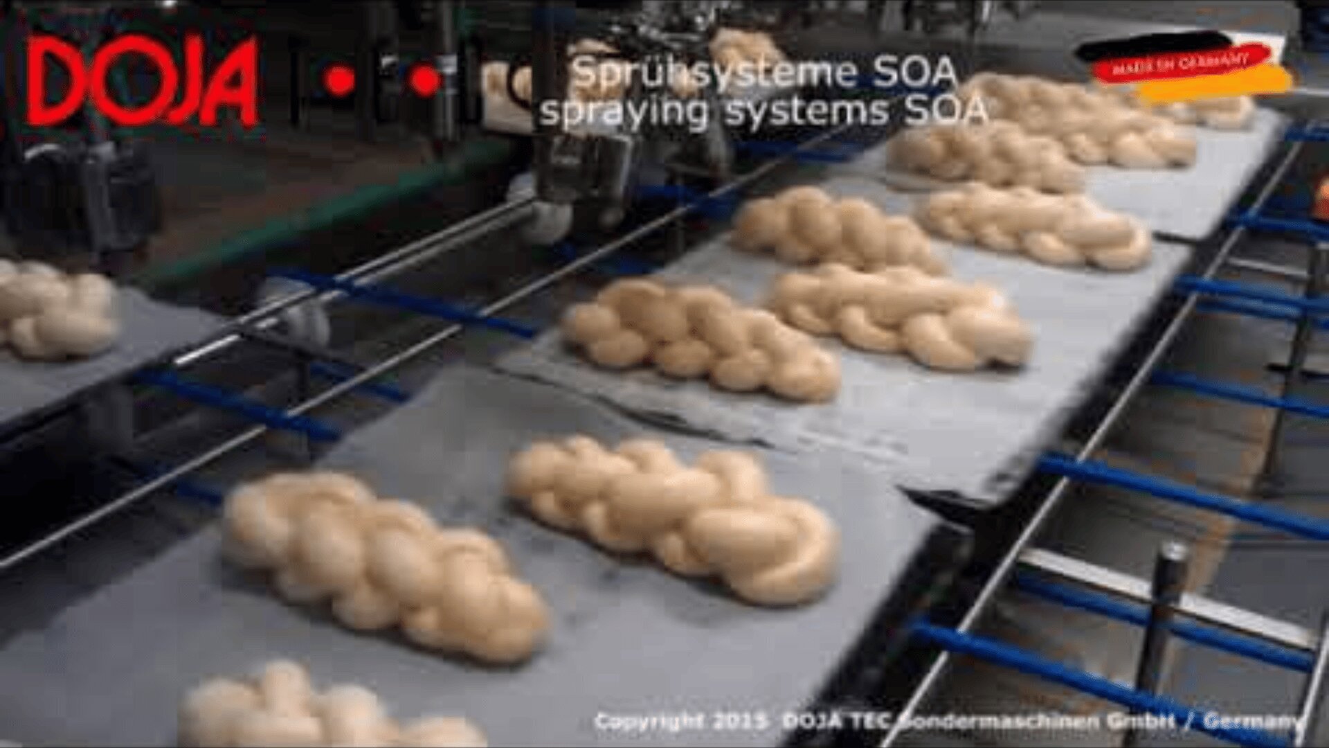 Backwaren laufen auf einem Förderband unter industriellen Sprühsytemen mit der Aufschrift "Sprühsyteme SOA". Das DOJA-Logo und das Etikett "Made in Germany" sind auf dem Bild zu sehen.