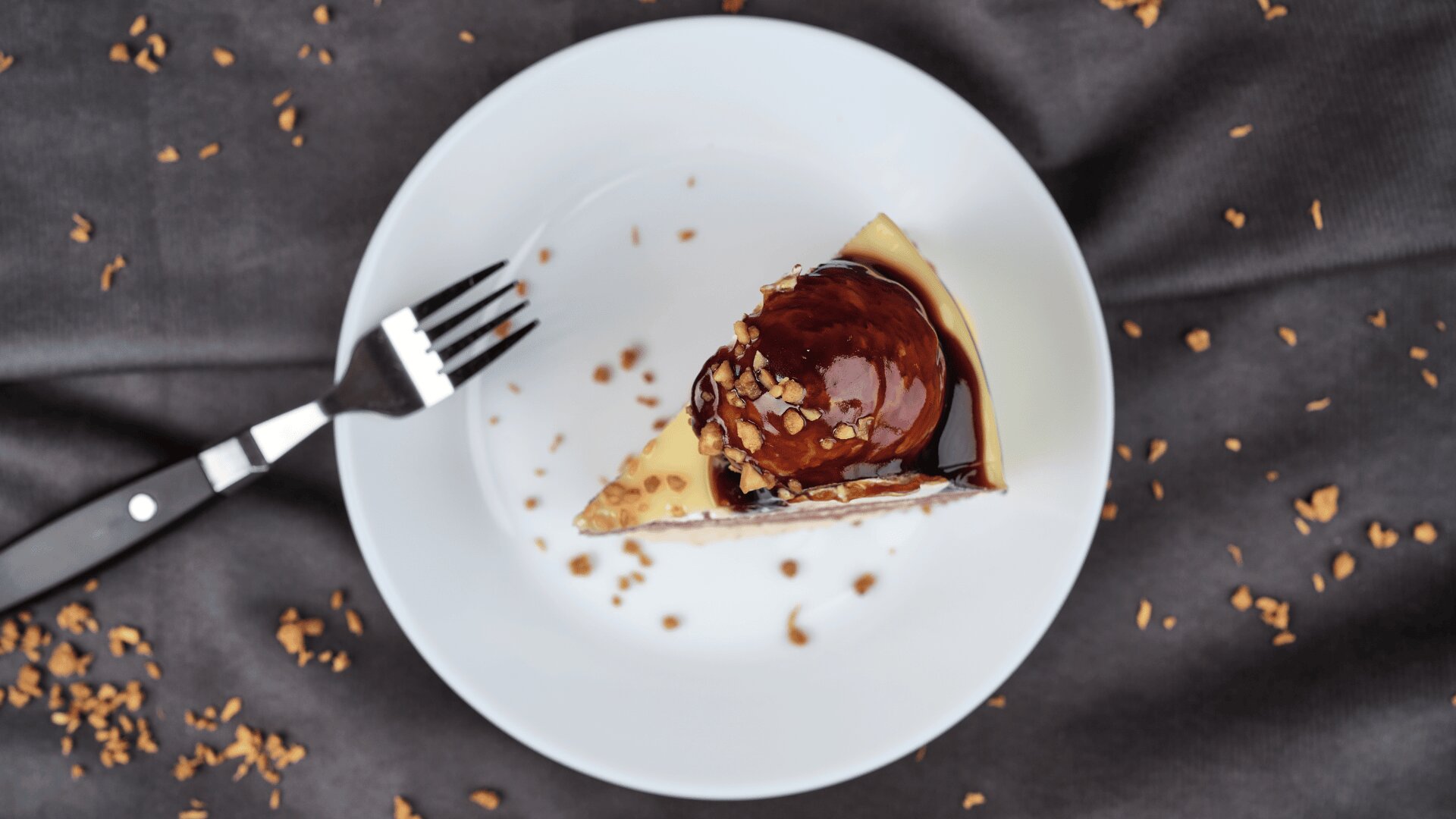 A slice of cheesecake topped with chocolate sauce and crushed nuts sits on a white plate with a fork beside it, all on a dark cloth background with scattered nut pieces.