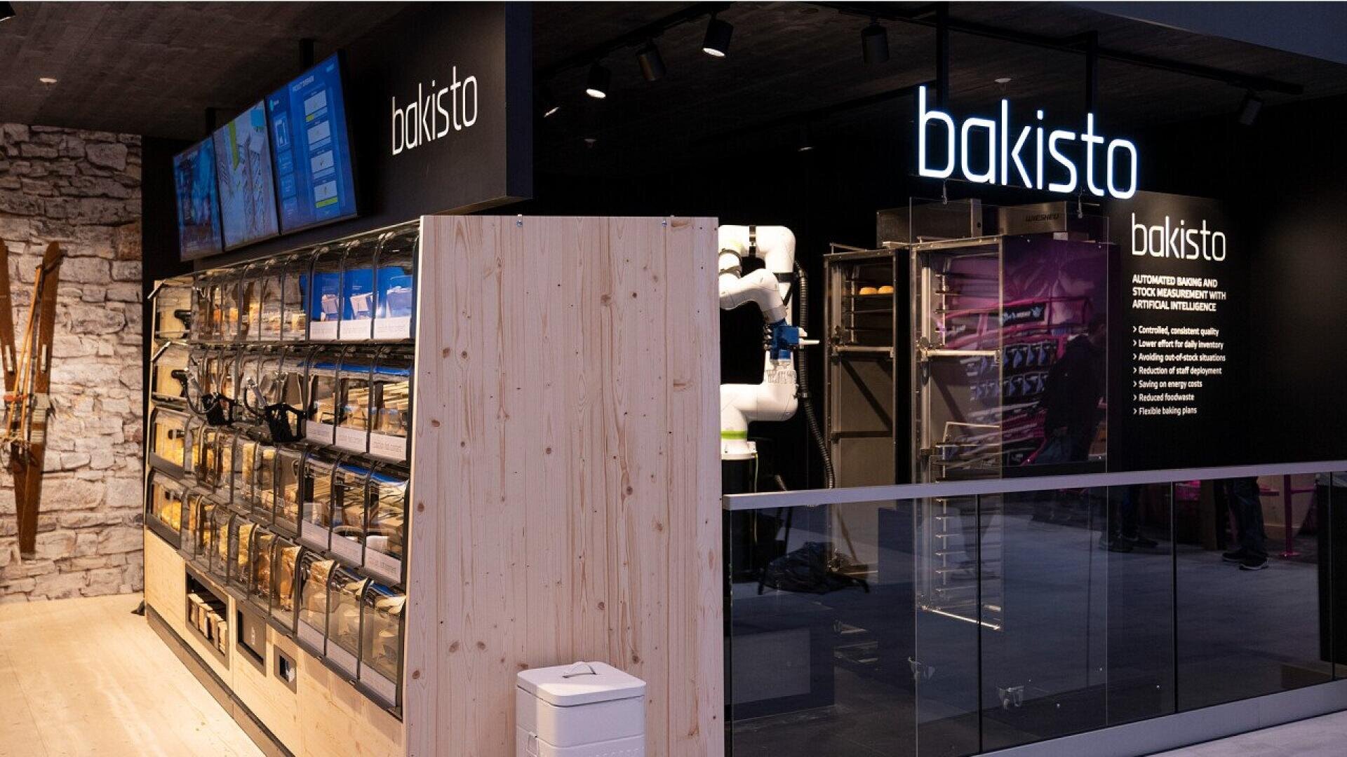 A modern ski rental shop with a wall of organized ski equipment, a digital display screen, and a sign that reads bakisto above a sleek, glass-enclosed equipment area.