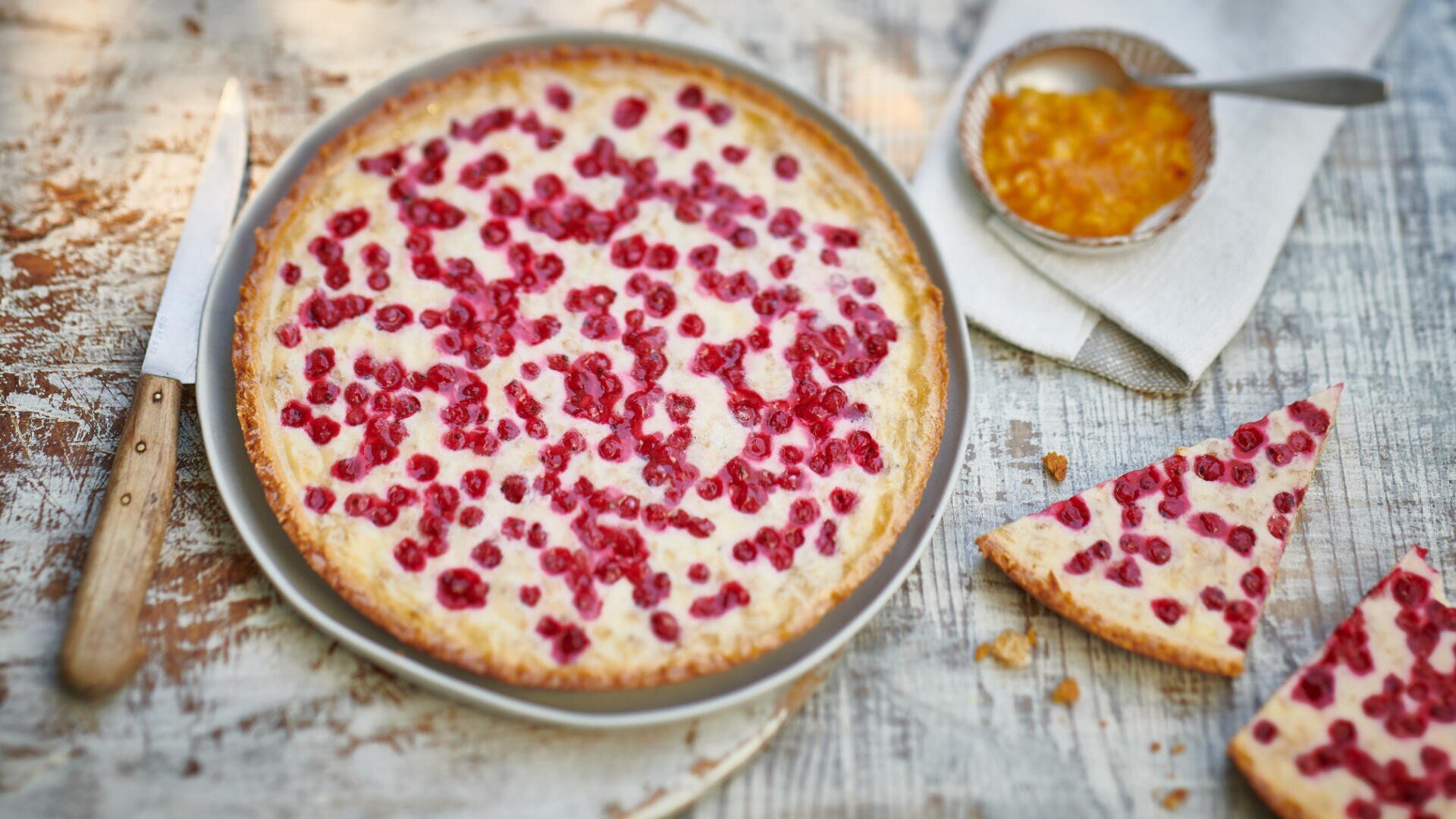 Eine runde Beerentorte mit goldener Kruste und roten Beeren obendrauf steht auf einem rustikalen Holztisch neben einem Messer, einer Schüssel Marmelade und zwei dreieckigen Tortenstücken auf einer Serviette.