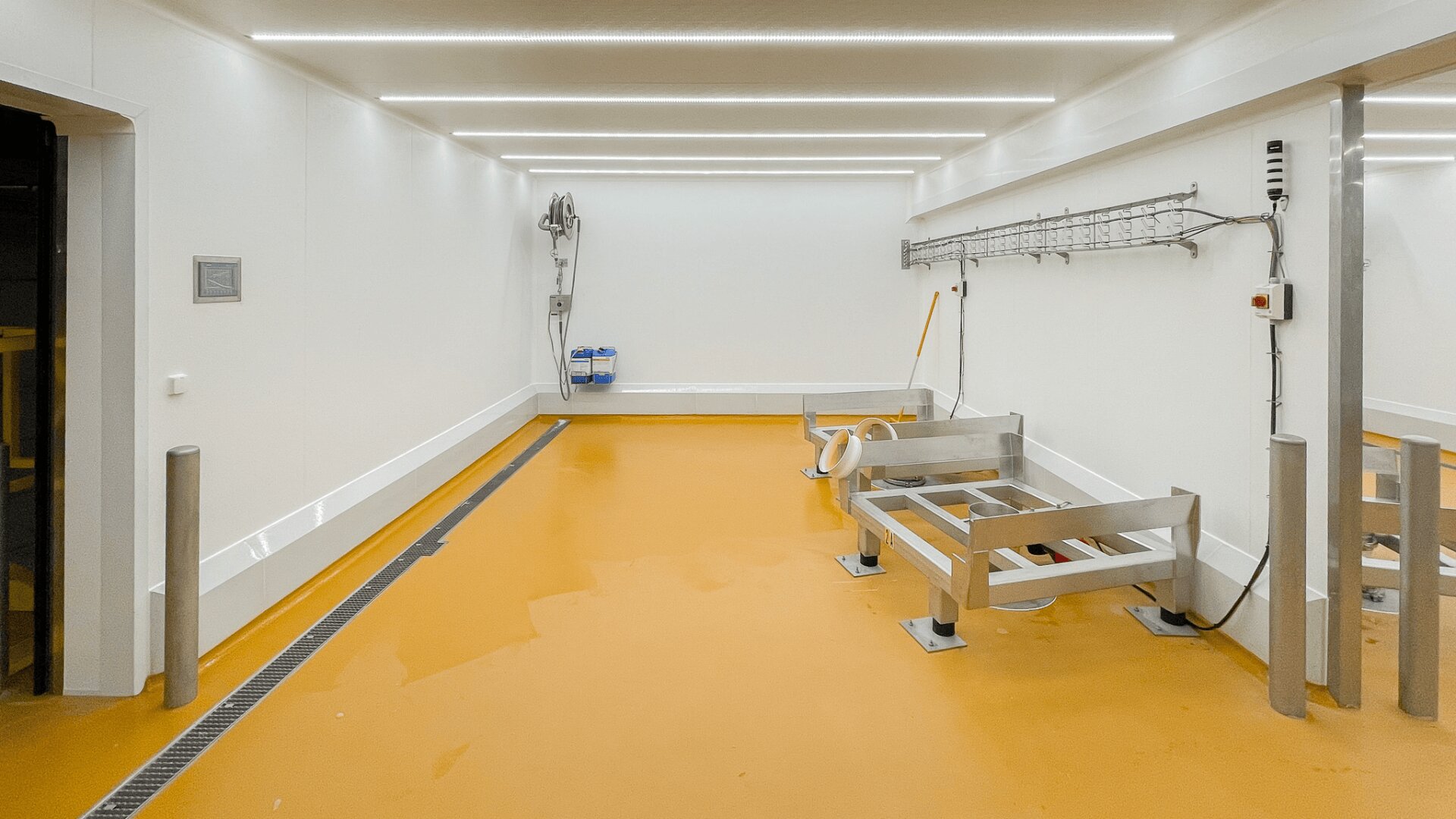 Ein sauberer, hell erleuchteter Raum mit weißen Wänden und orangefarbenem Boden, in dem Metallregale, ein Schlauch, eine Wandspule und Haken an einer Wand angebracht sind. Der Raum wirkt industriell und gut gepflegt.