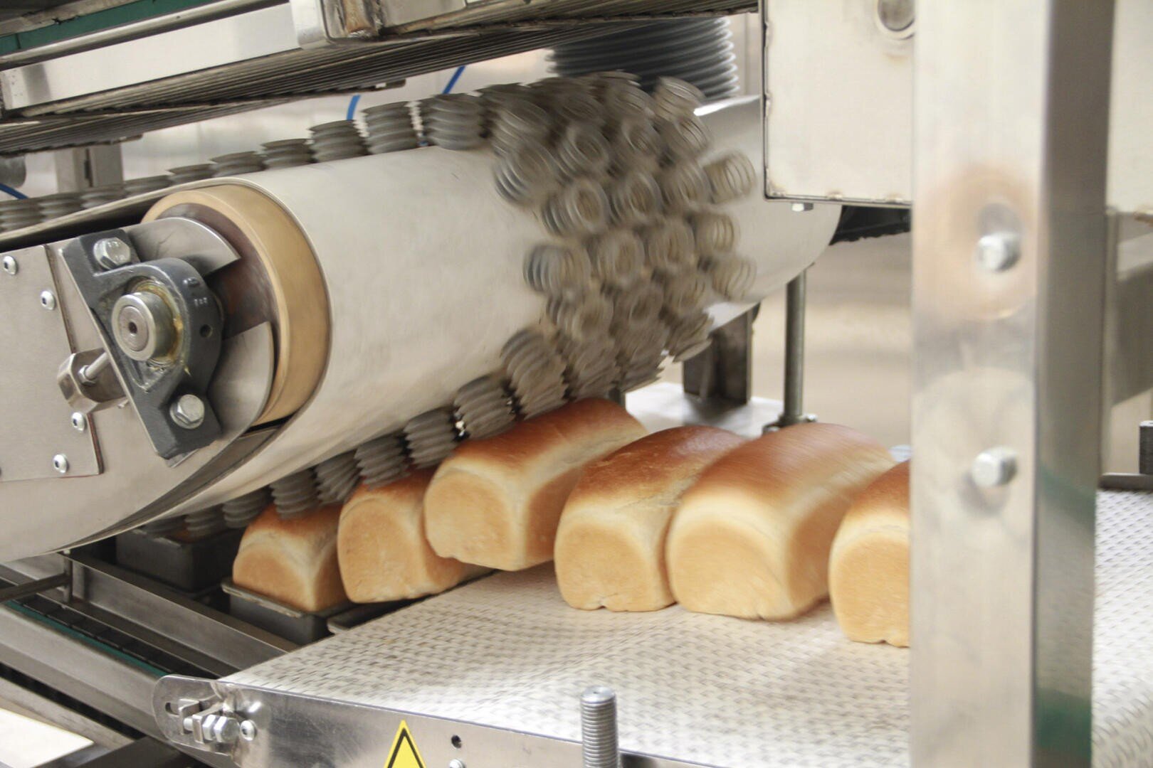 Brotlaibe bewegen sich auf einem Förderband unter einer Maschine mit strukturierten Rollen in einer Brotfabrik, was auf einen automatisierten Brotproduktionsprozess hindeutet.