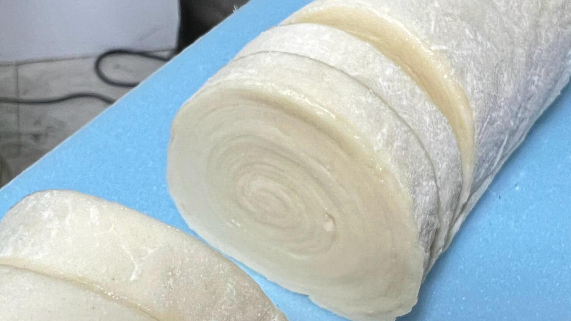 A close-up of a rolled dough log that has been sliced into thick, round pieces on a blue cutting board. The dough has a spiral pattern and a lightly floured surface.