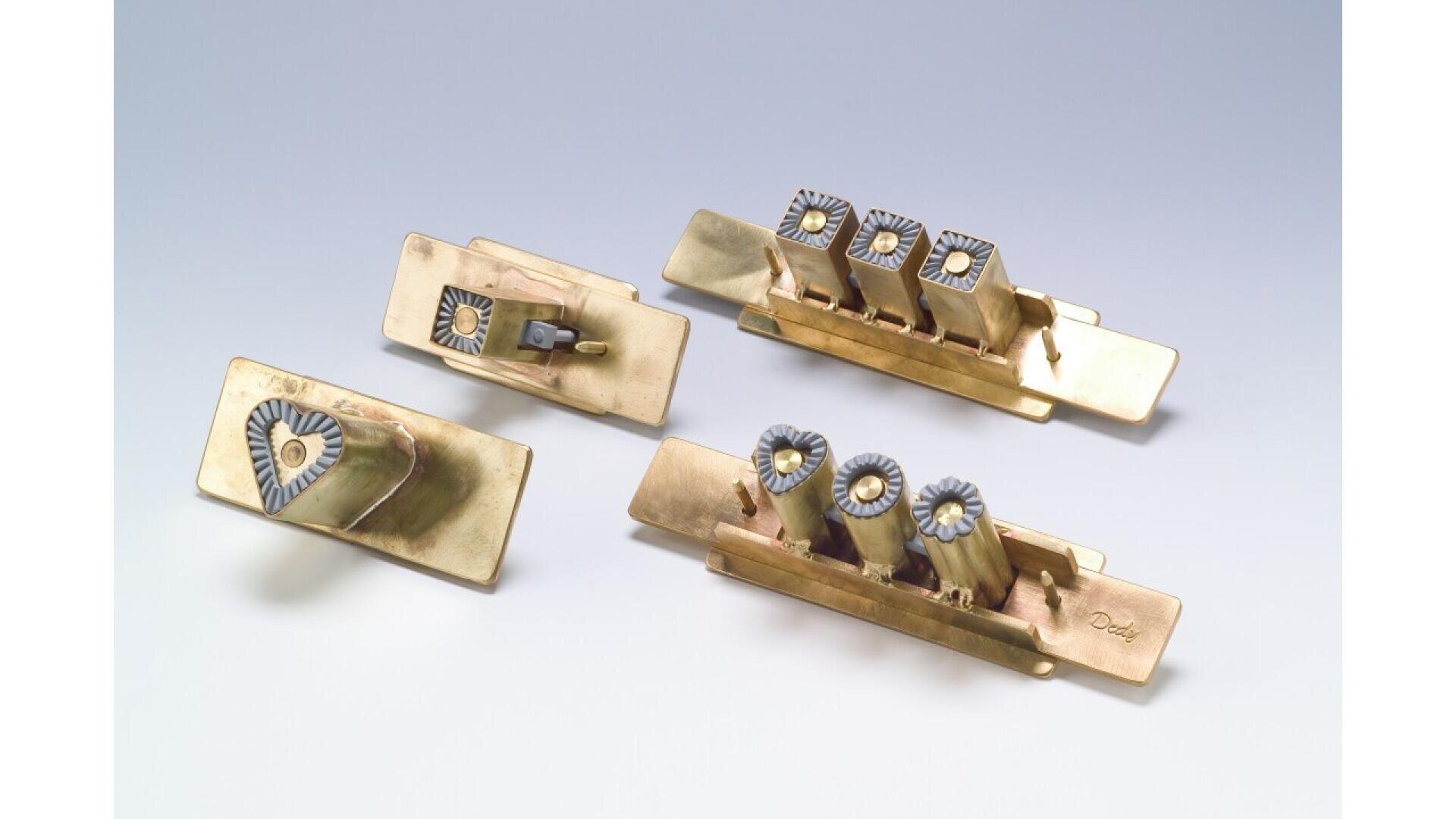 Three brass branding irons with rectangular bases and handles, each featuring raised shapes or letters on the end for imprinting designs. The items are arranged on a light, neutral background.