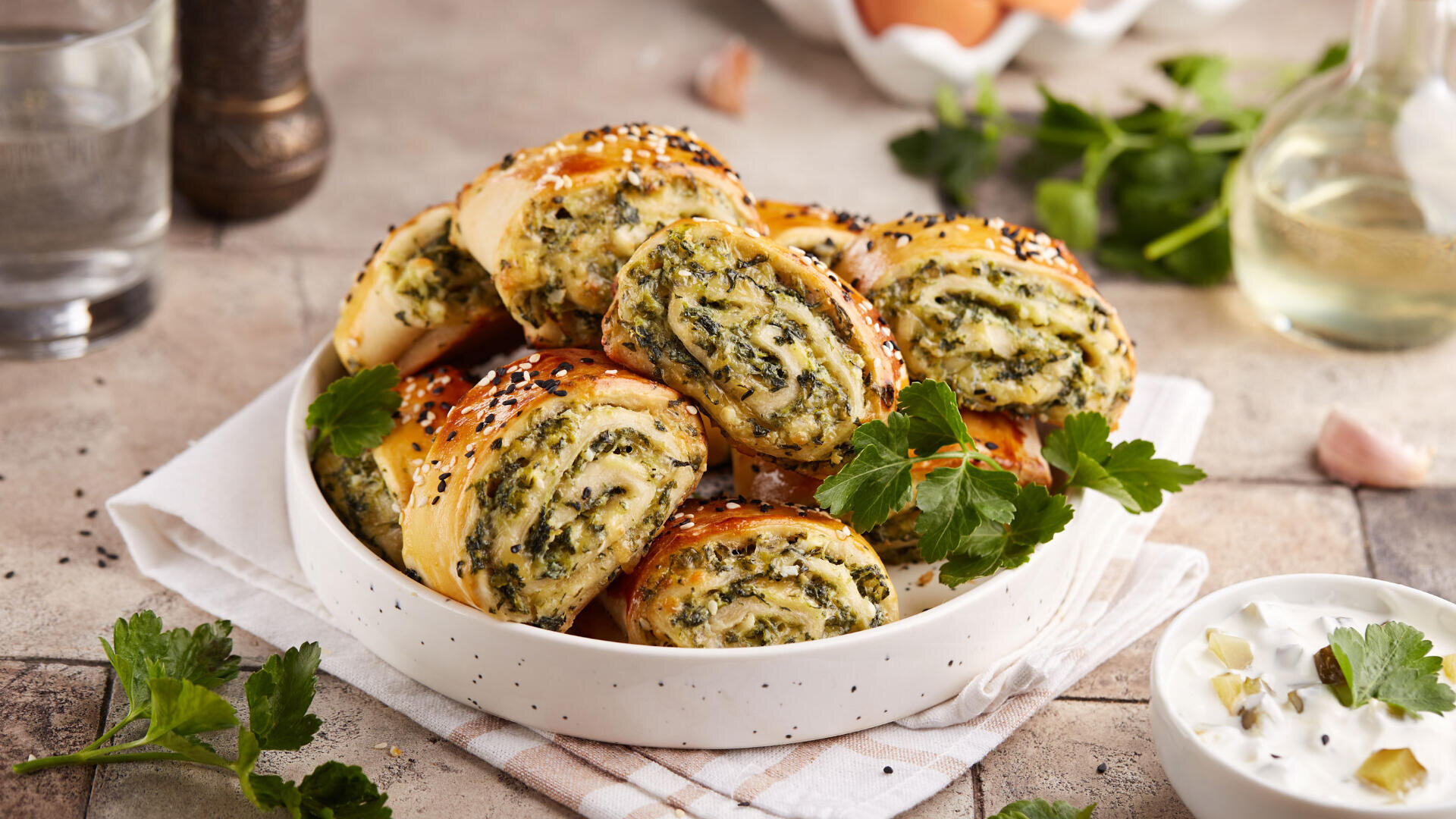 Auf einer Stoffserviette steht eine Schale mit Scheiben von herzhaften Teigrollen, gefüllt mit Spinat und Käse, garniert mit Sesam. Frische Petersilie und eine Schale mit cremigem Dip stehen daneben.