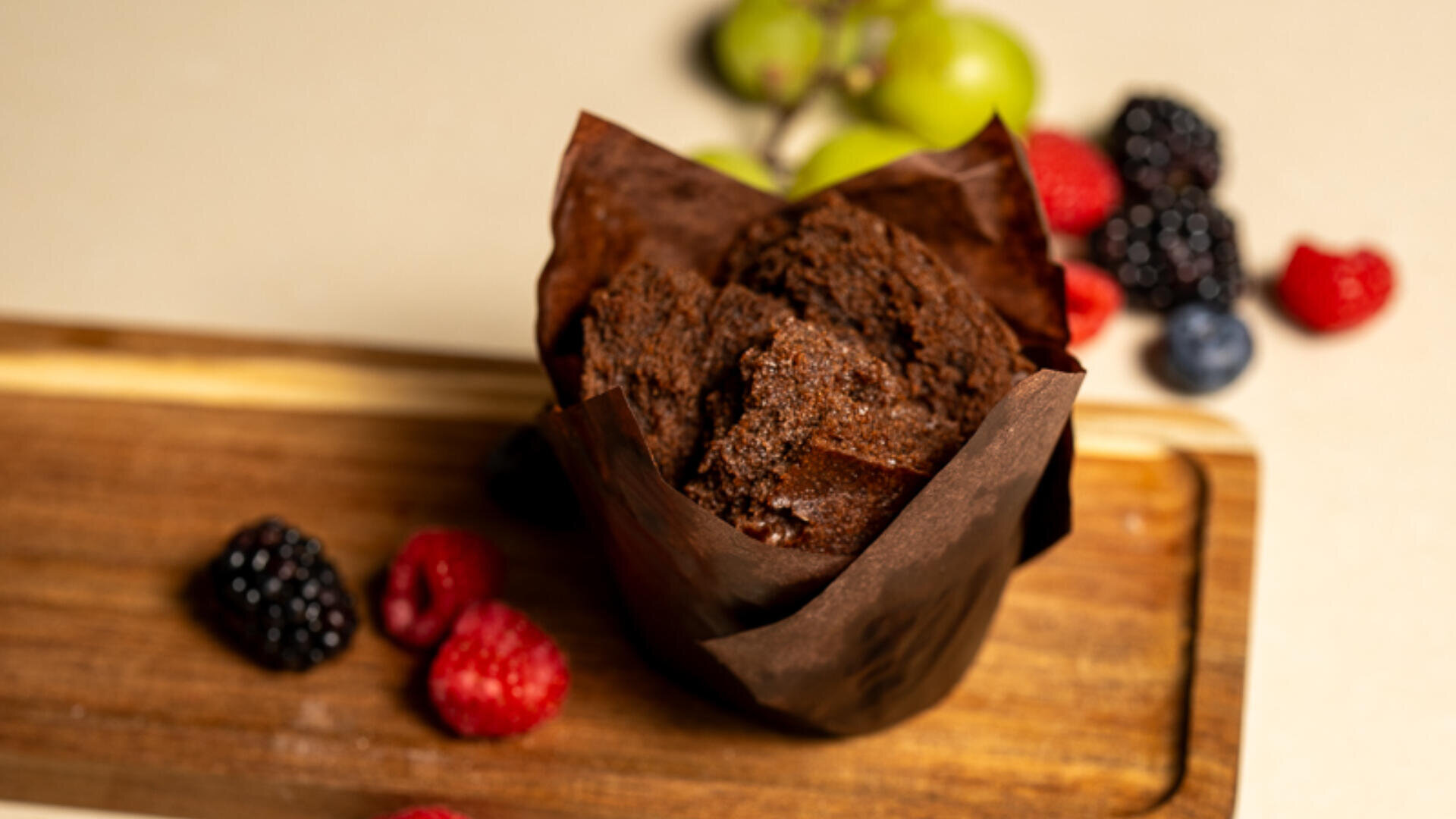 A chocolate muffin in brown paper sits on a wooden board, surrounded by fresh raspberries, blackberries, blueberries, and green grapes in the background.