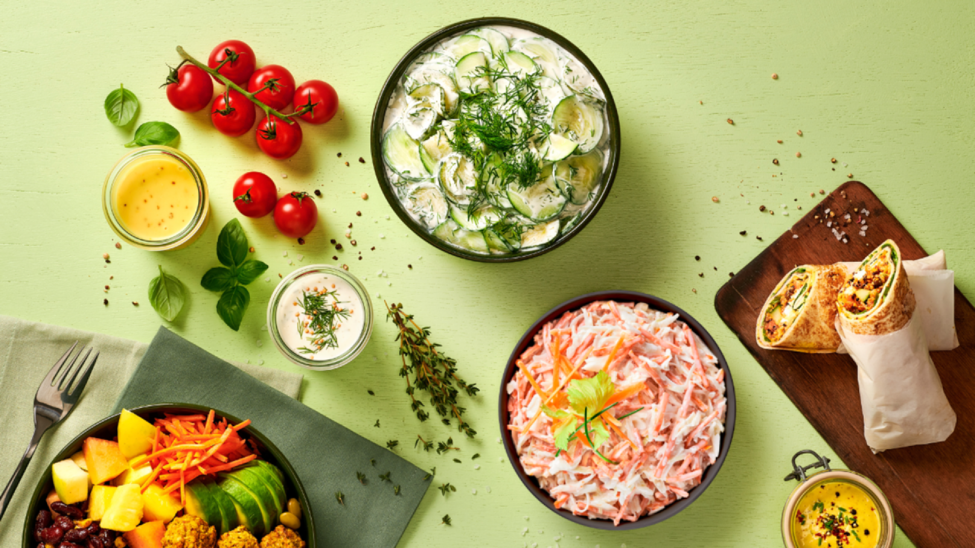 A colorful spread of fresh salads, cherry tomatoes, herbs, salad dressings, a wrap cut in halves, and a bowl of mixed fruit and falafel arranged on a light green table.