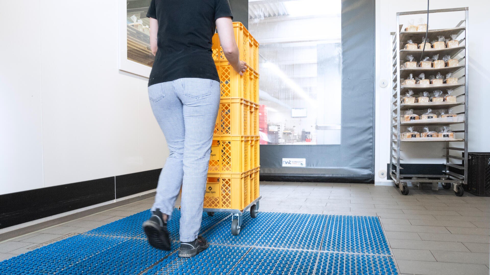 Eine Person in karierten Hosen schiebt einen mit gelben Kisten beladenen Wagen über eine blaue Matte in einer sauberen Industrieumgebung; im Hintergrund sind Regale mit Behältern zu sehen.