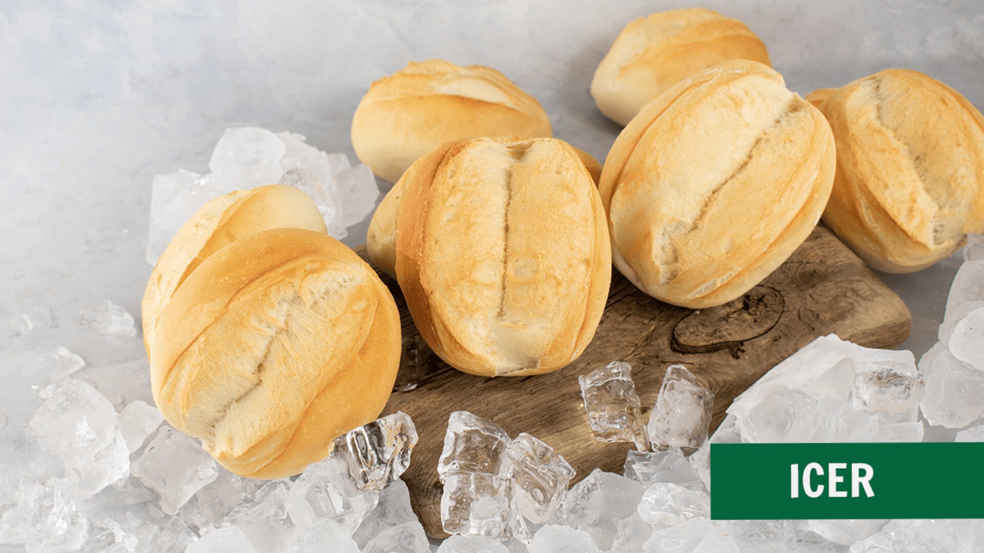 Several crusty bread rolls are placed on a wooden board surrounded by ice cubes on a light surface. A green box in the lower right corner displays the word “ICER.”.
