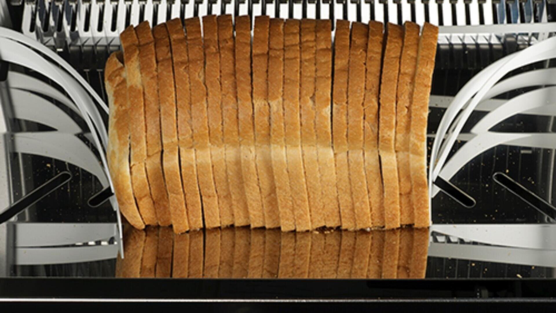 A loaf of bread is being evenly sliced by an industrial bread-slicing machine, with metal blades separating each slice.