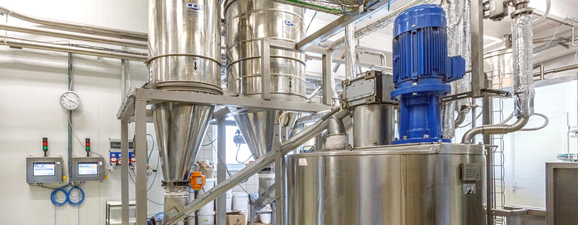 Industrial facility with shiny metal tanks, pipes, and machinery, including a large blue motor. Two wall-mounted control panels and a clock are visible, all in a clean, bright environment.