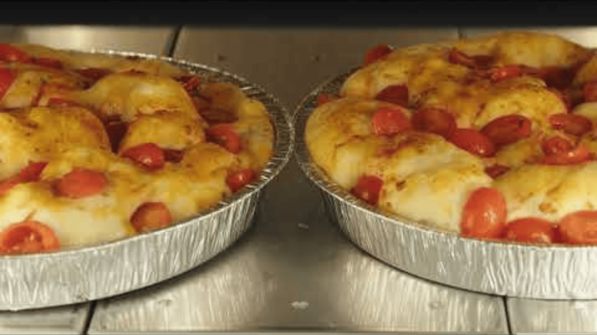 Two round foil pans filled with baked bread, topped with melted cheese and sliced cherry tomatoes, side by side on a rack in the oven.