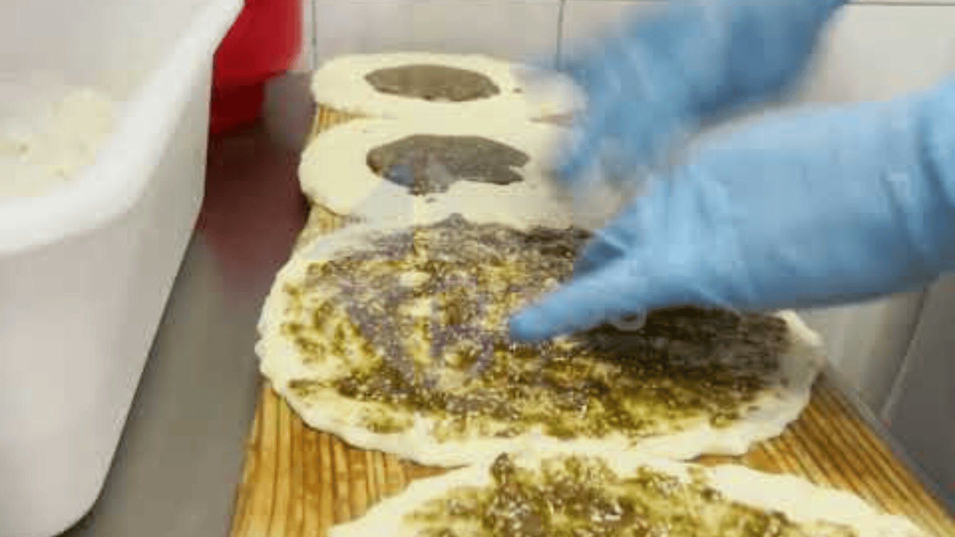 A person wearing blue gloves spreads a topping onto flat rounds of dough on a wooden board, preparing multiple pieces for baking. A white container with ingredients is visible beside the dough.