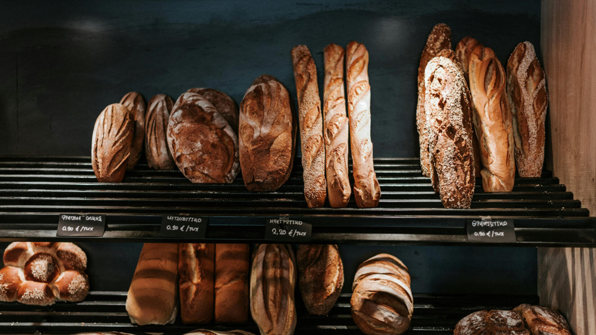Eine Vielzahl von handwerklich hergestellten Broten und Baguettes, die in einer Bäckerei auf Metallregalen ausgestellt sind, mit handgeschriebenen Etiketten vor jeder Sorte.