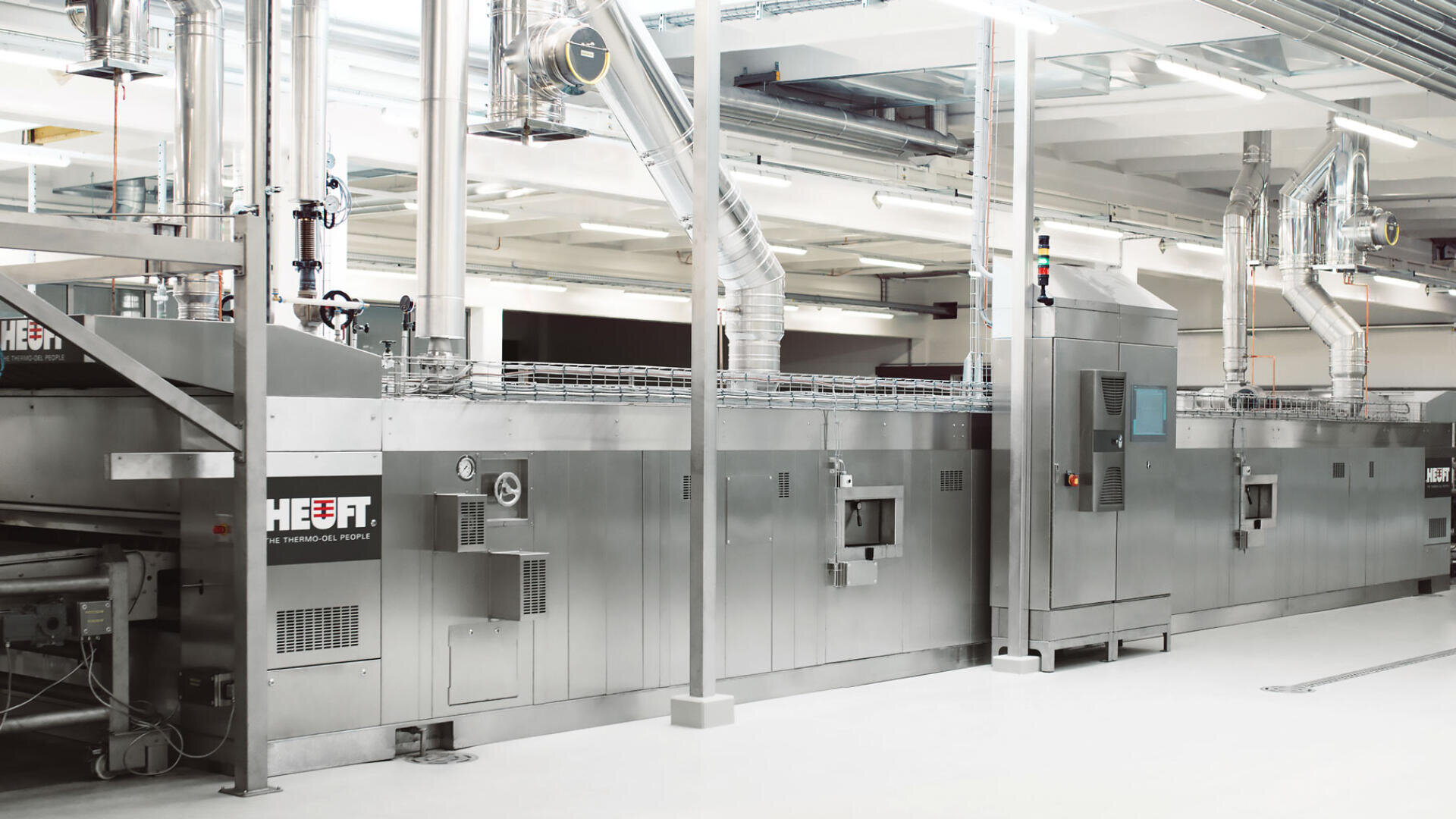 Eine große Industriemaschine aus rostfreiem Stahl mit Entlüftungsöffnungen, Rohren und Bedienfeldern in einer sauberen, hell erleuchteten Fabrikumgebung. Die Maschine ist mit "HEUFT." beschriftet.