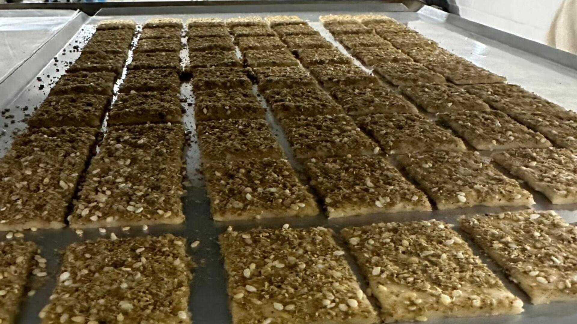 Reihen von rechteckigen Fladenbroten, die mit einer Mischung aus Sesamsamen und Gewürzen belegt sind, werden auf einem Metallblech zum Backen bereitgelegt.