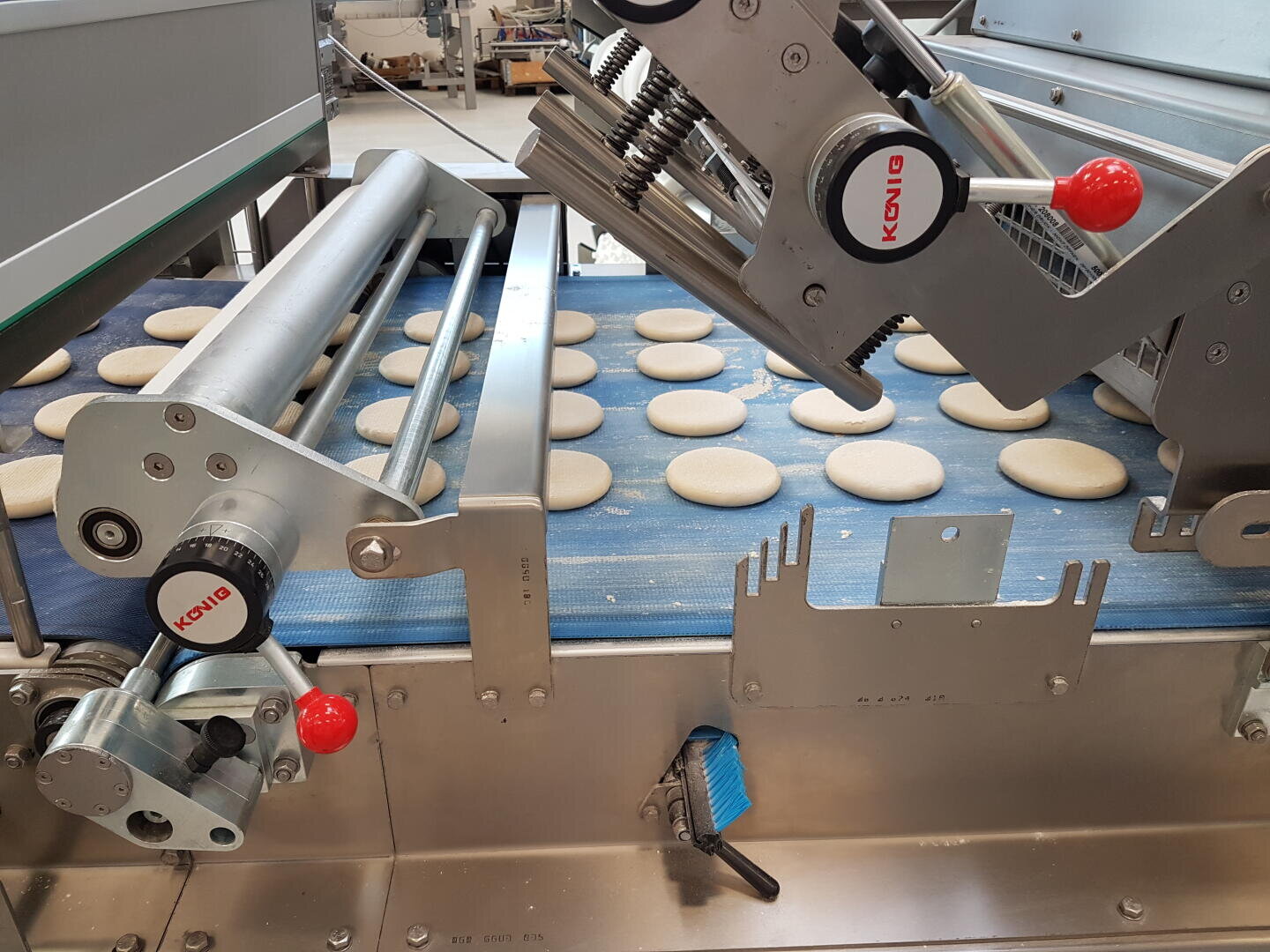 Round dough pieces are evenly spaced on a blue conveyor belt inside an industrial baking machine labeled KONIG, with metal parts and red handles visible.