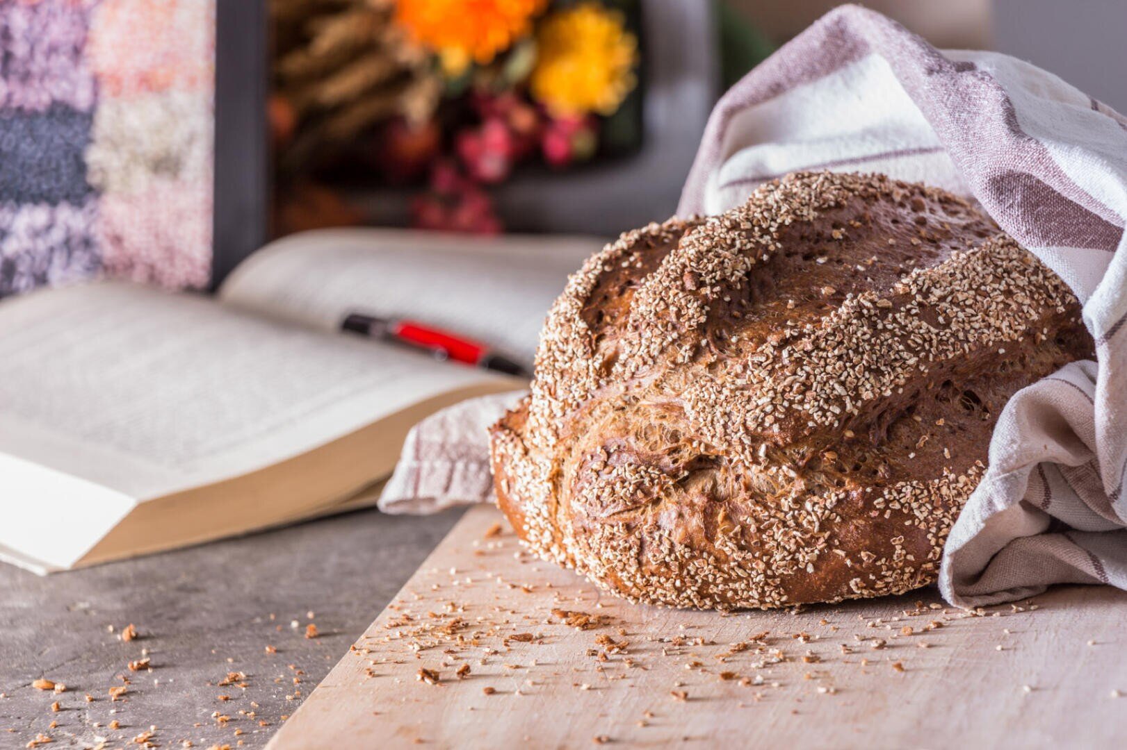 A loaf of seeded bread partially covered with a cloth sits on a wooden board, surrounded by crumbs. An open book and pen are blurred in the background, along with a bouquet of flowers.
