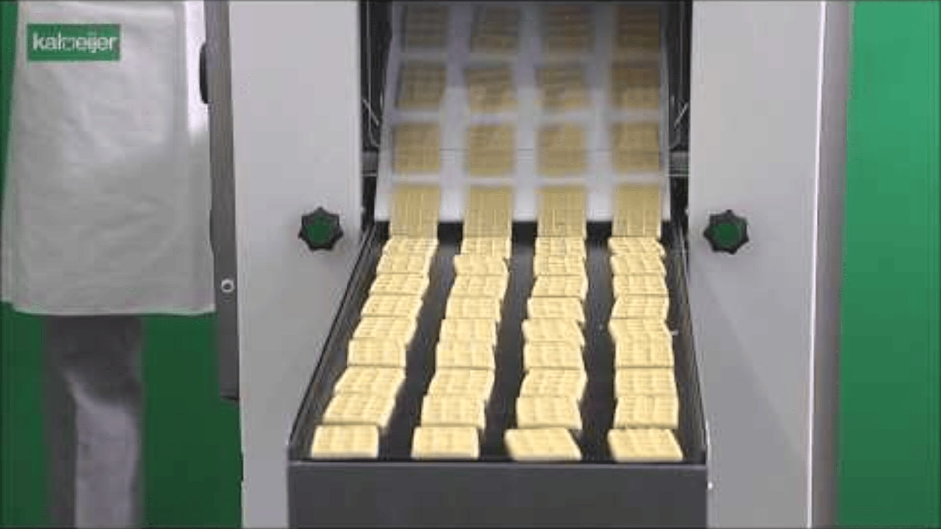 Eine Maschine produziert Reihen von rechteckigen Crackern oder Keksen auf einem Förderband. Auf der linken Seite steht eine Person in einem weißen Laborkittel. Der Hintergrund ist grün, und der Markenname Kalterer erscheint in der oberen linken Ecke.