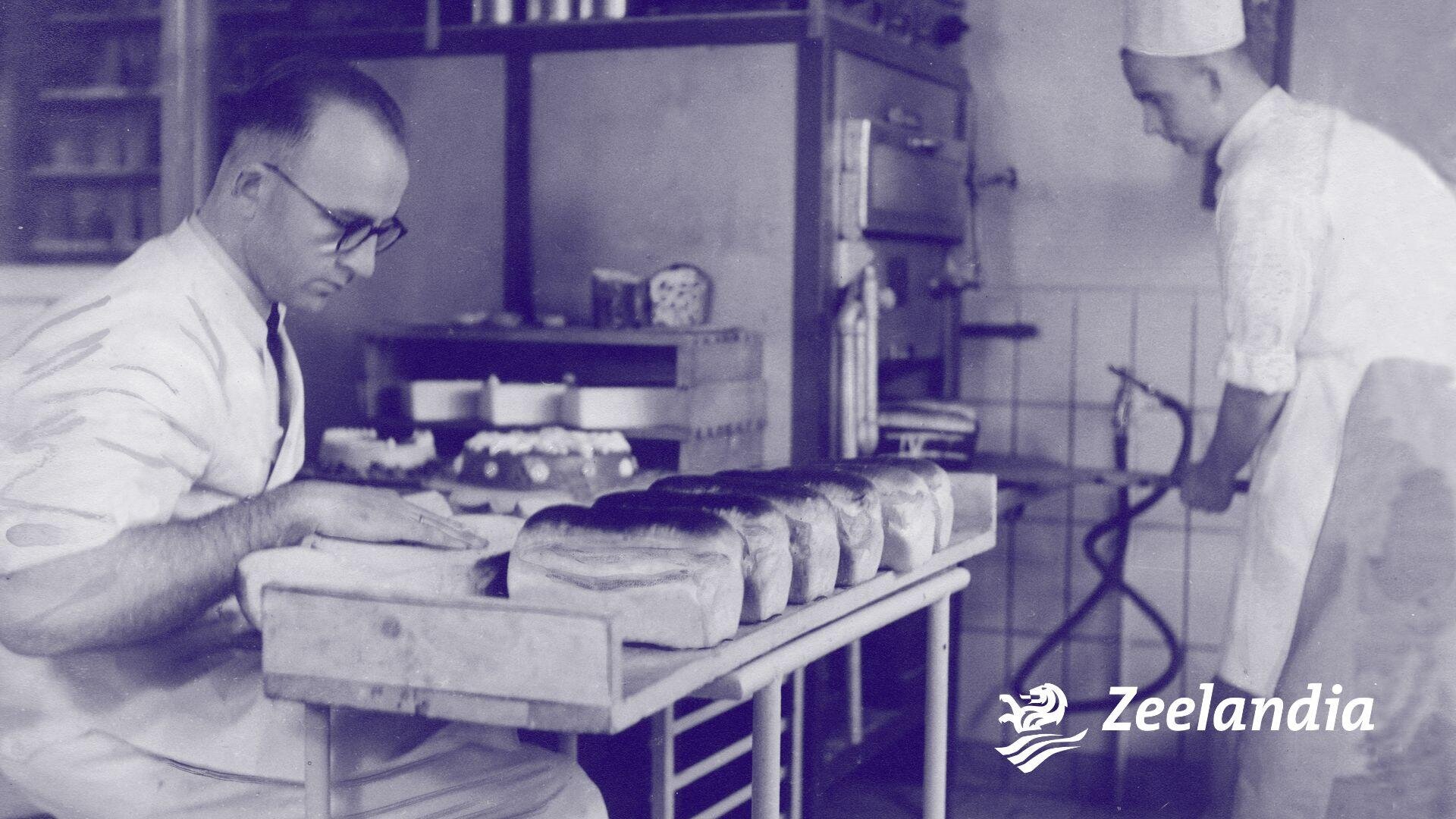 Two bakers work in a kitchen; one inspects loaves of bread on a table while the other uses an oven. The room has baking equipment and trays. The Zeelandia logo is in the bottom right corner.