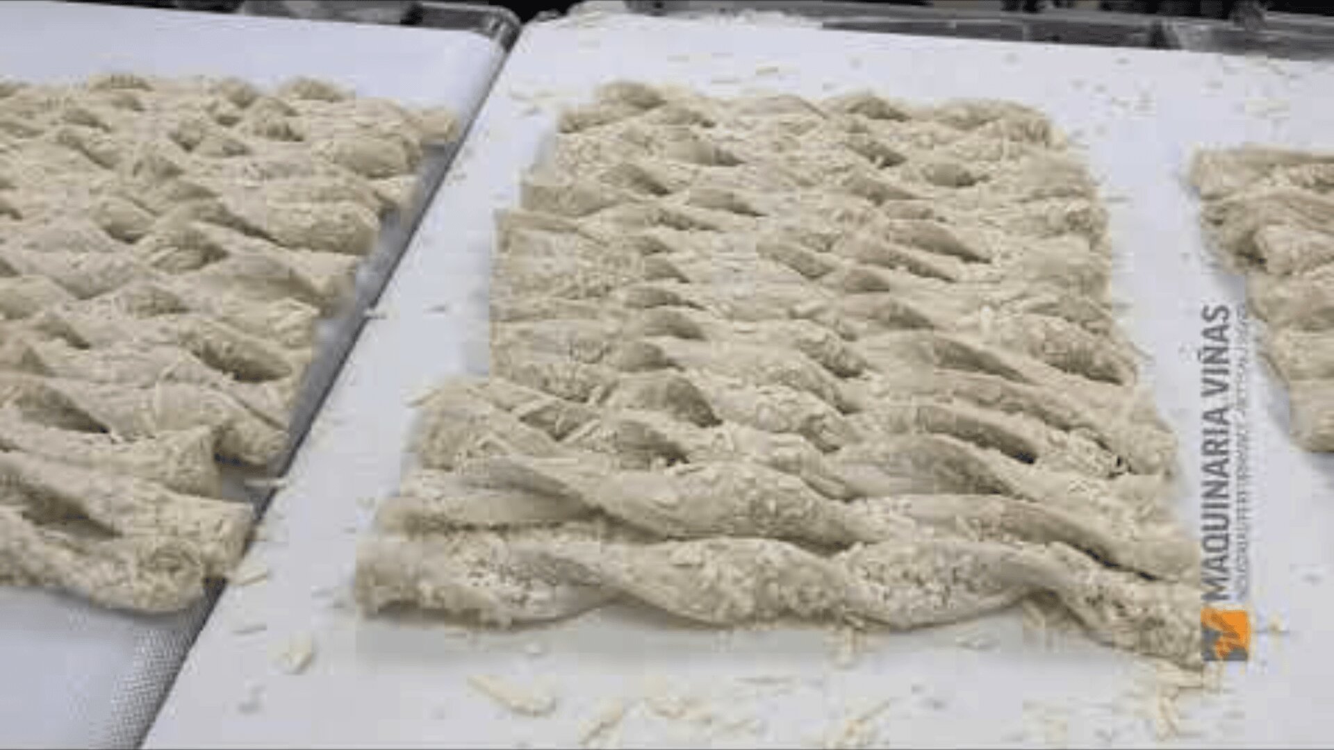 Rows of raw, braided dough sheets with a lattice pattern are placed on a conveyor, ready for baking. The surface is dusted with flour, and the logo MAQUINARIA VIÑAS is visible on the right side.