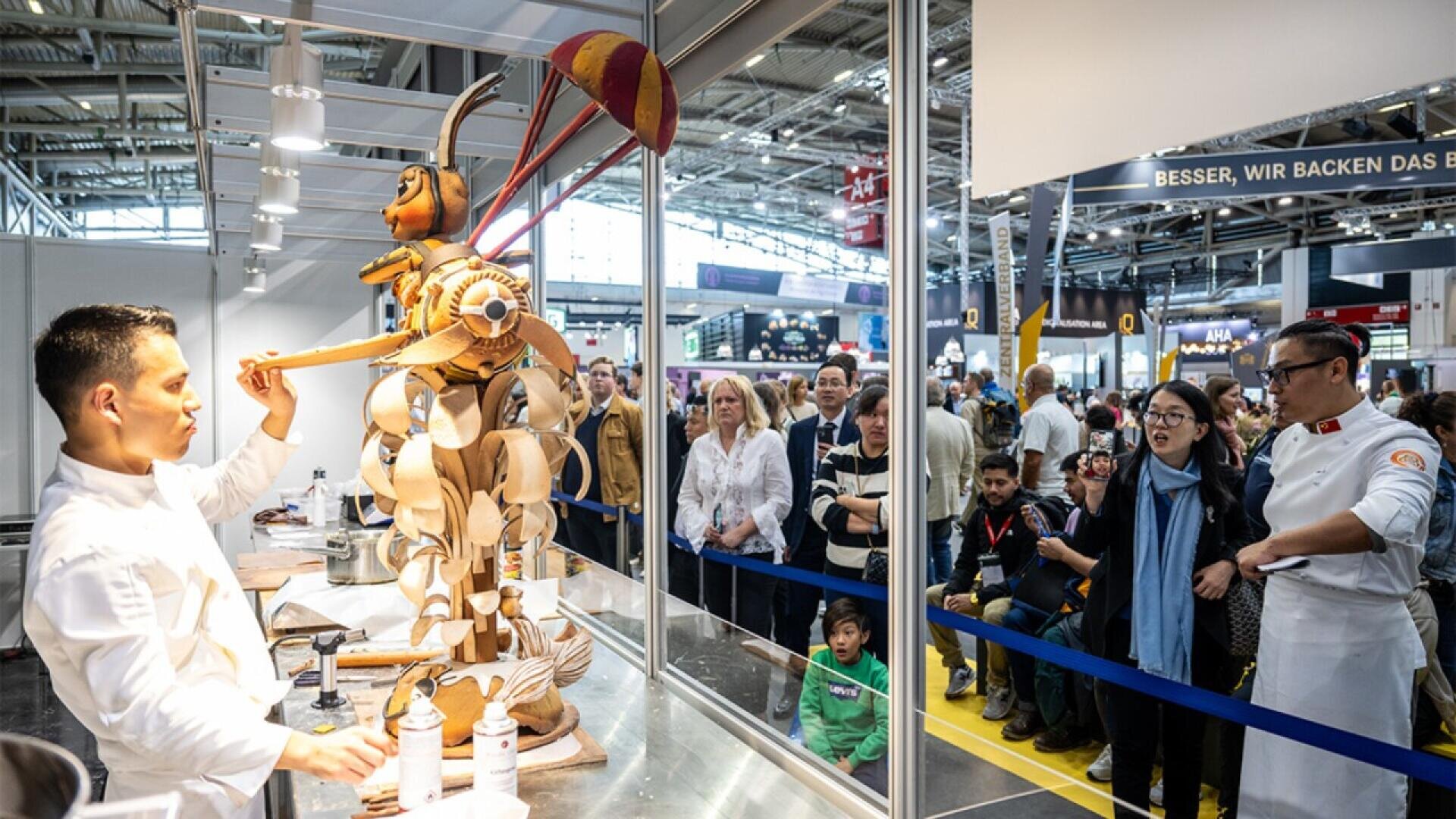 A chef presents a large, intricate pastry sculpture of an insect with an umbrella to a crowd of onlookers at an indoor event, while another chef stands nearby. The crowd watches with interest behind a blue barrier.