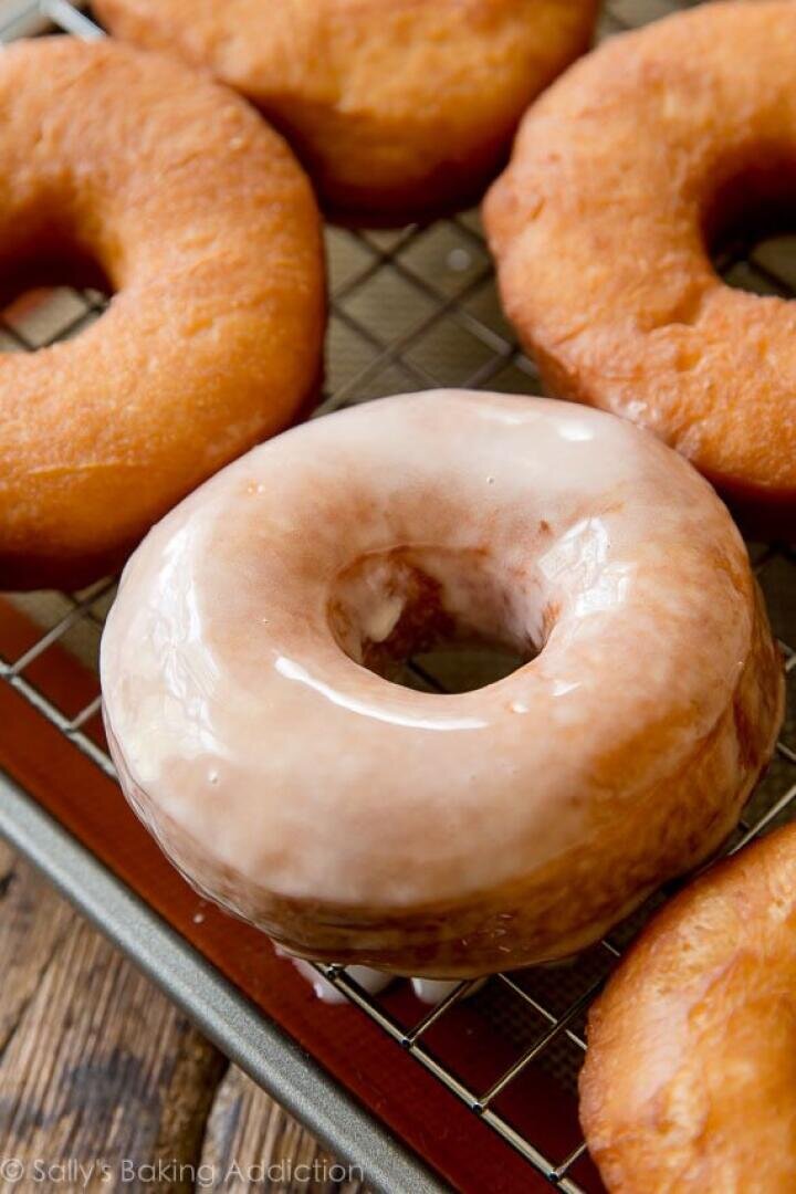 A close-up of several homemade donuts on a cooling rack, with one donut in the foreground covered in a shiny glaze while the others remain plain.