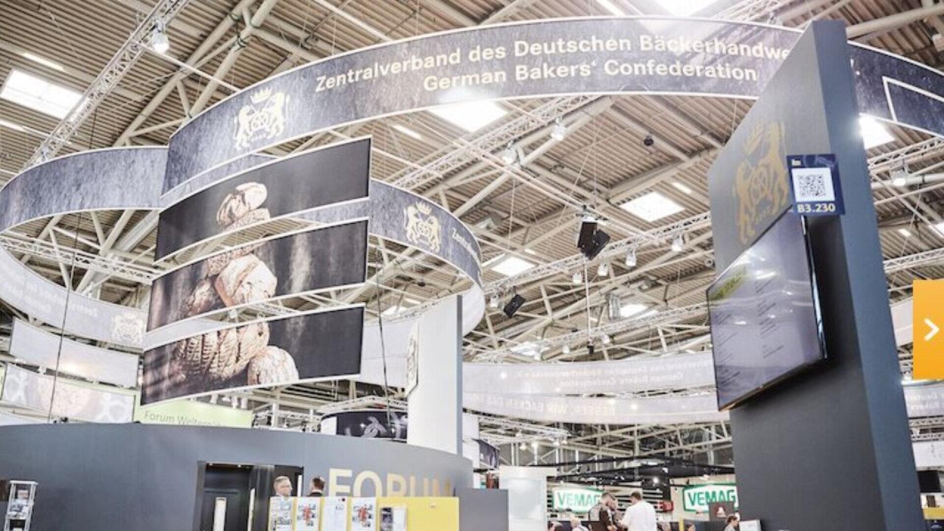 A large exhibition booth for the German Bakers’ Confederation features images of bread, digital screens, and a prominent sign in German and English inside a spacious, modern convention center.