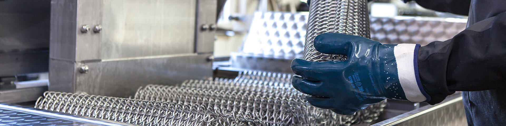 Eine Person mit blauen Schutzhandschuhen handhabt einen zylindrischen Metallgewebefilter in der Nähe von Industrieanlagen aus Edelstahl.