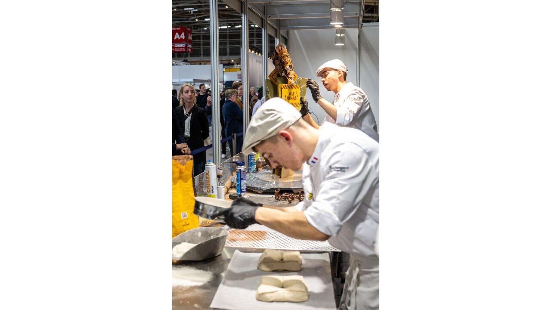 Two bakers in white uniforms and hats work in a bright, busy bakery. One focuses on shaping dough on a tray, while the other decorates a tall pastry. People watch them through a glass partition in the background.