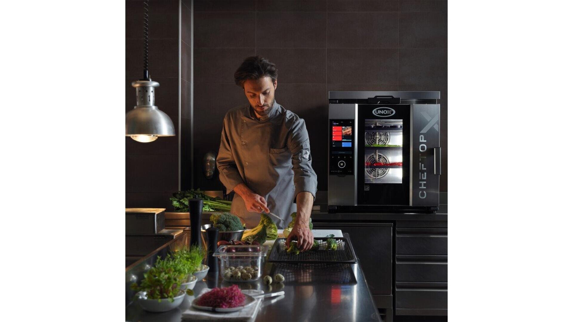 A chef in a modern kitchen prepares vegetables on a stainless steel counter, with fresh ingredients around him and a digital commercial oven in the background.