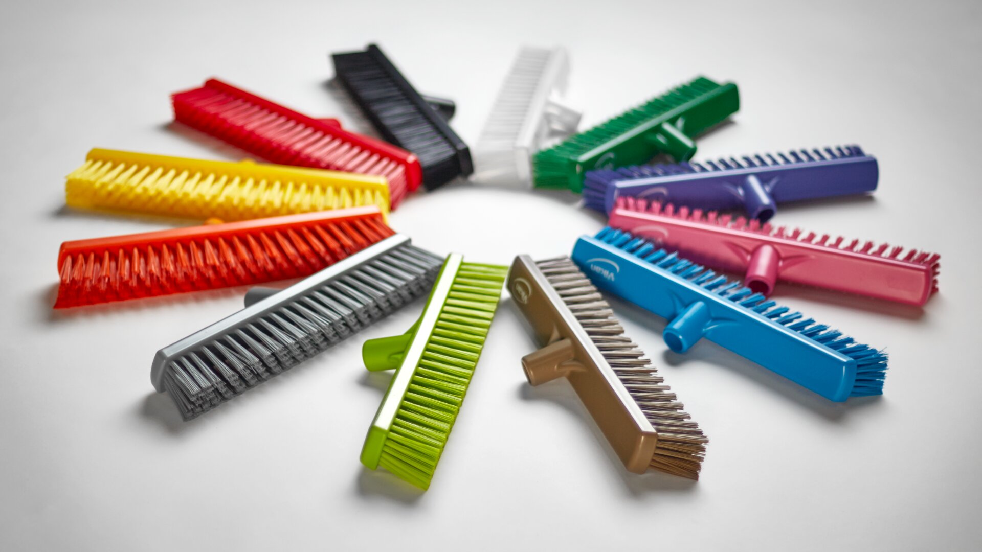A circle of colorful scrub brushes with bristles facing outward, arranged on a white surface. Each brush has a different colored handle, including red, yellow, green, blue, pink, black, and more.