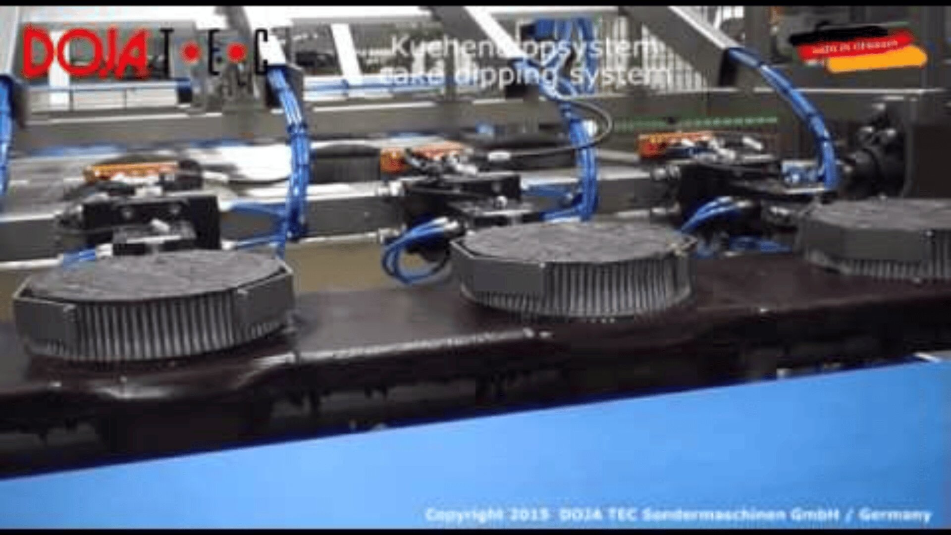 Automated cake dipping system with round cakes on a conveyor belt, blue hoses, and mechanical arms. Text on image includes “DOJA TEC,” “cake dipping system,” and company information at the bottom.