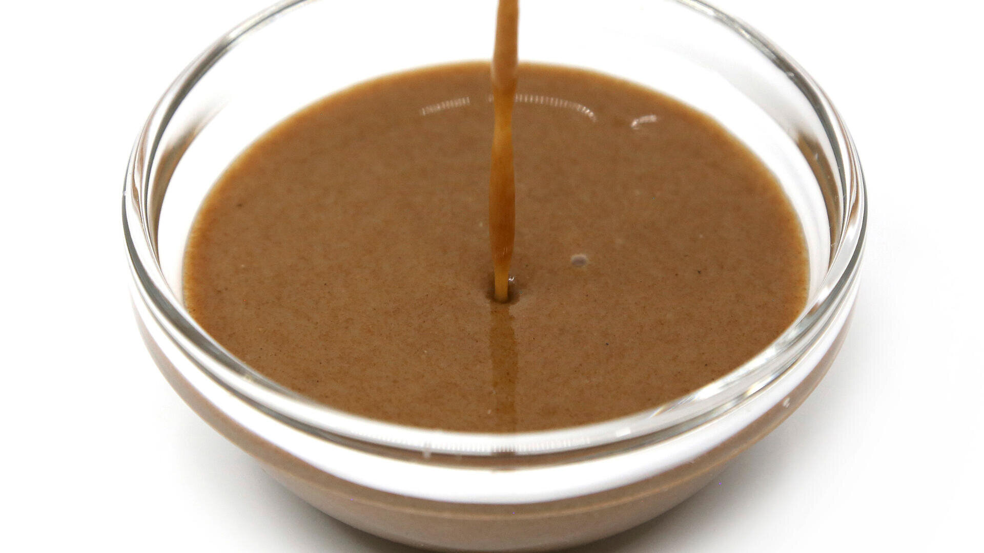 Ein Strahl brauner Tahinisauce wird in eine klare Glasschale gegossen, die mit mehr Sauce gefüllt ist, vor einem weißen Hintergrund.