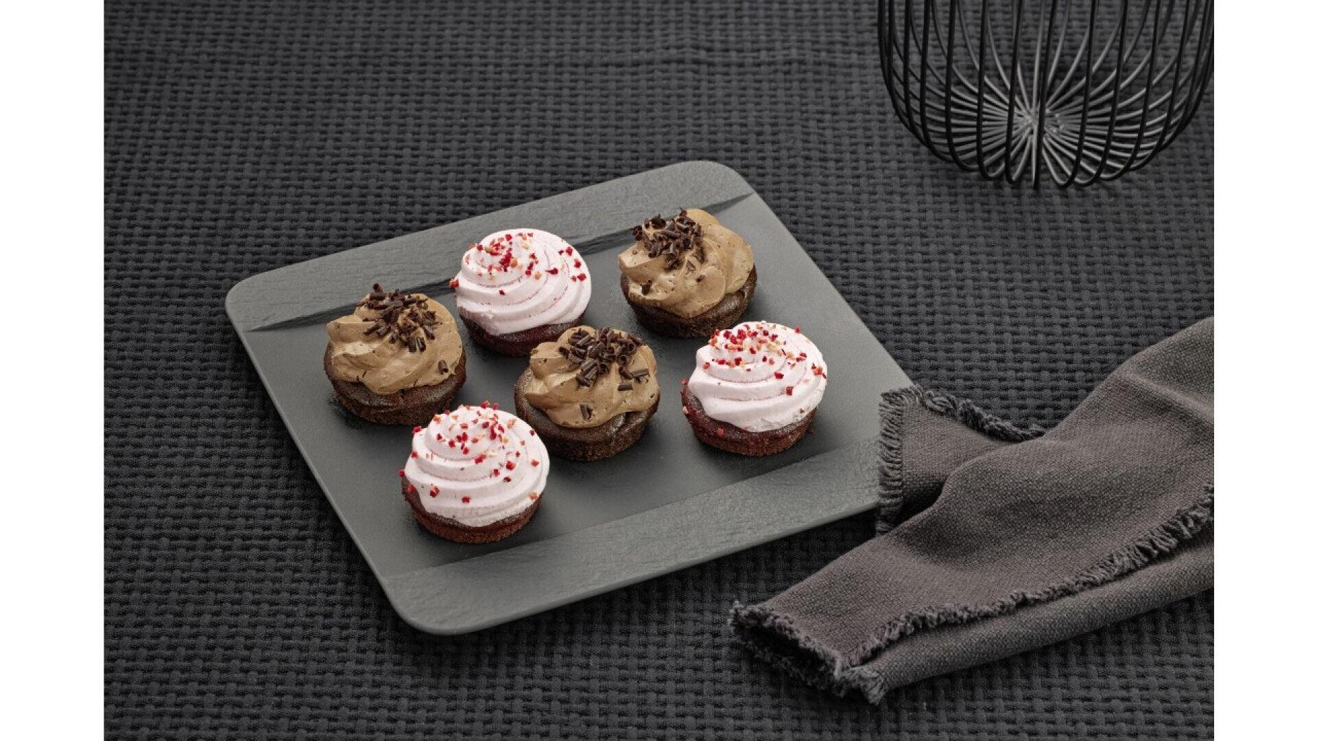 Six cupcakes topped with chocolate or pink frosting and sprinkles are arranged on a square slate tray, set on a textured black surface beside a dark cloth napkin and a decorative black wire object.