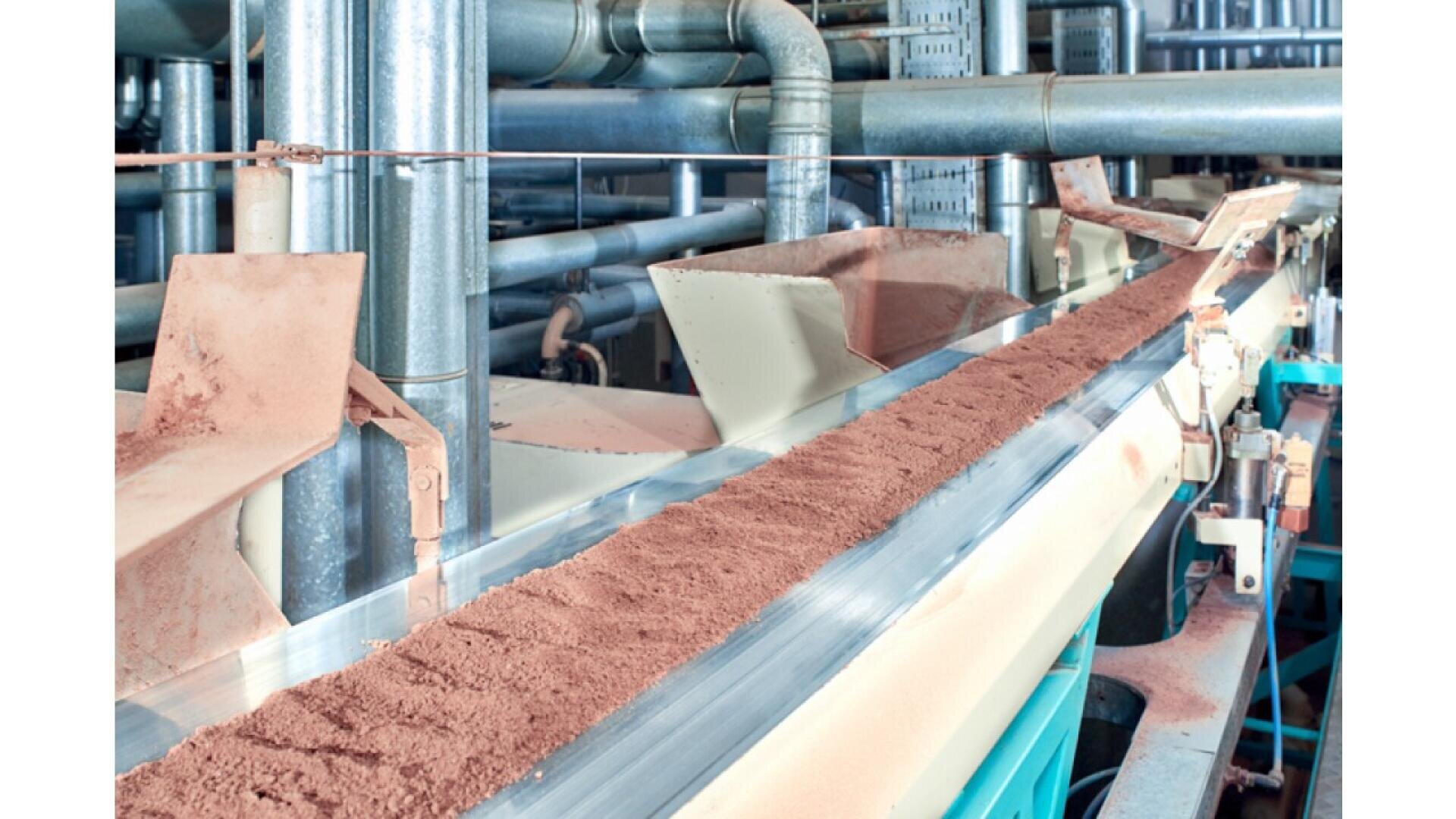 A conveyor belt transports a continuous stream of reddish-brown powder through an industrial facility with metal pipes and machinery in the background.