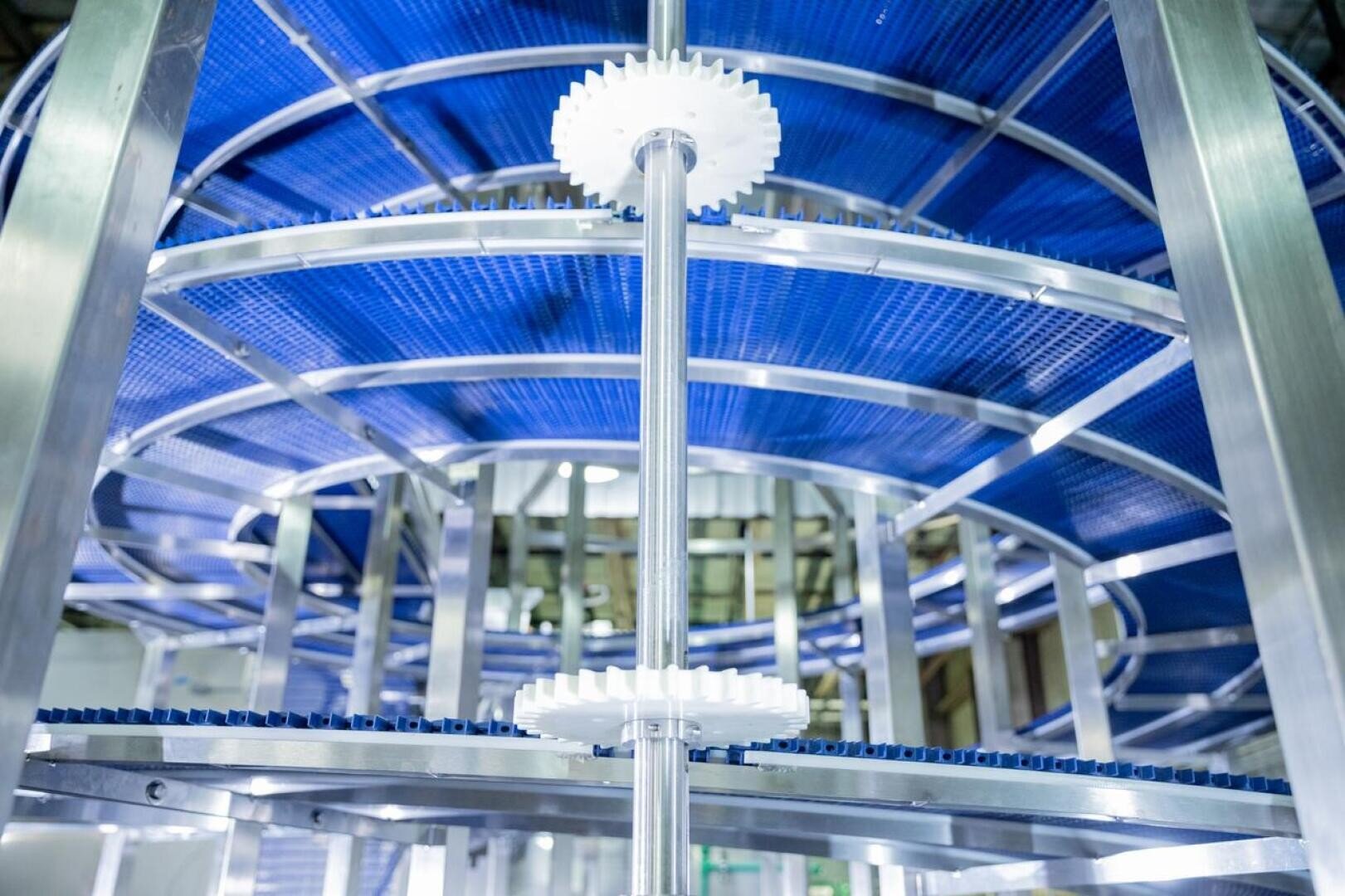 Close-up view of a spiral conveyor system with blue belts and white plastic gears inside a modern industrial facility. The metallic structure and circular design are prominent.