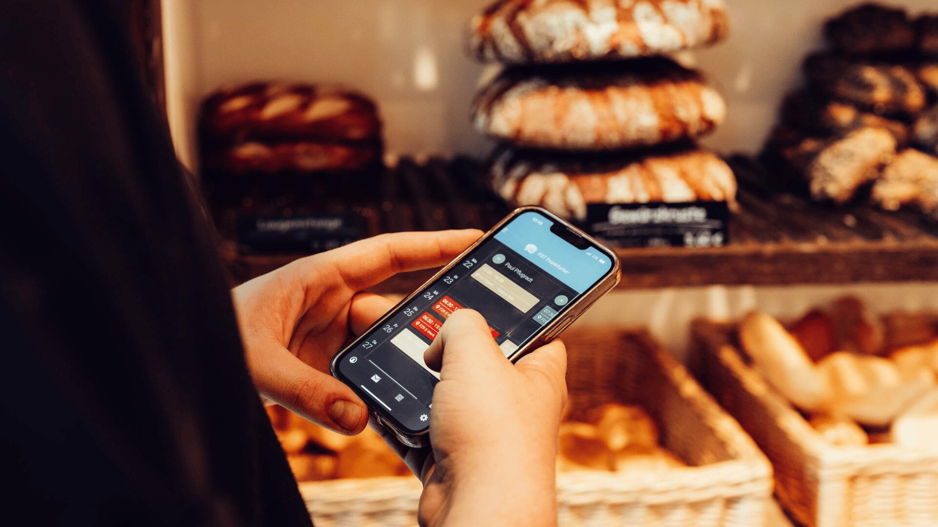 Eine Person benutzt ein Smartphone in einer Bäckerei, während im Hintergrund Regale mit verschiedenen Brotsorten und Gebäck zu sehen sind.