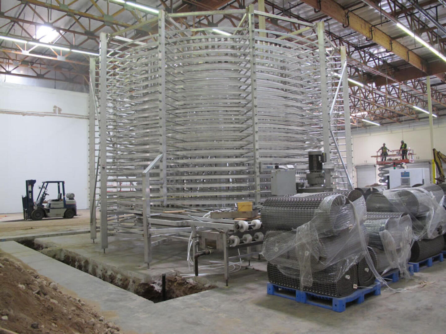 Large industrial spiral conveyor system being installed in a spacious warehouse, with a forklift and other equipment nearby. Some conveyor components are wrapped in plastic and placed on blue pallets.