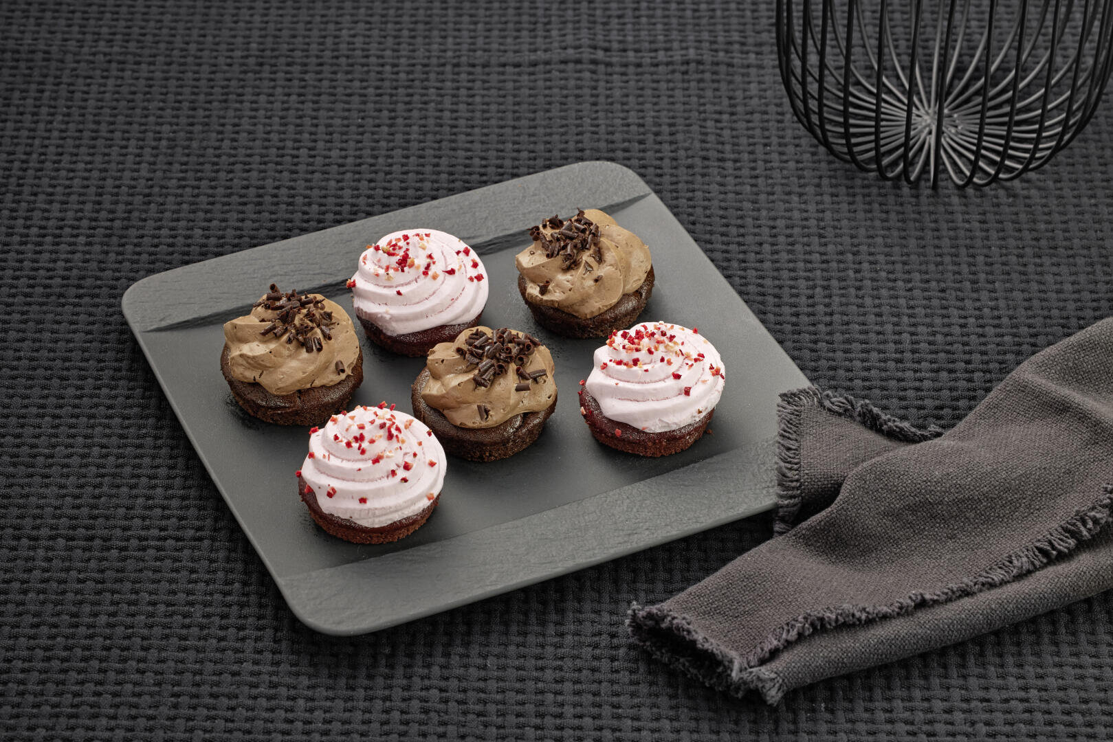 Sechs Cupcakes mit rosafarbenem und braunem Zuckerguss, einige mit roten Streuseln und andere mit Schokoladensplittern, sind auf einer Schieferplatte neben einem grauen Tuch auf einer strukturierten schwarzen Oberfläche angeordnet.