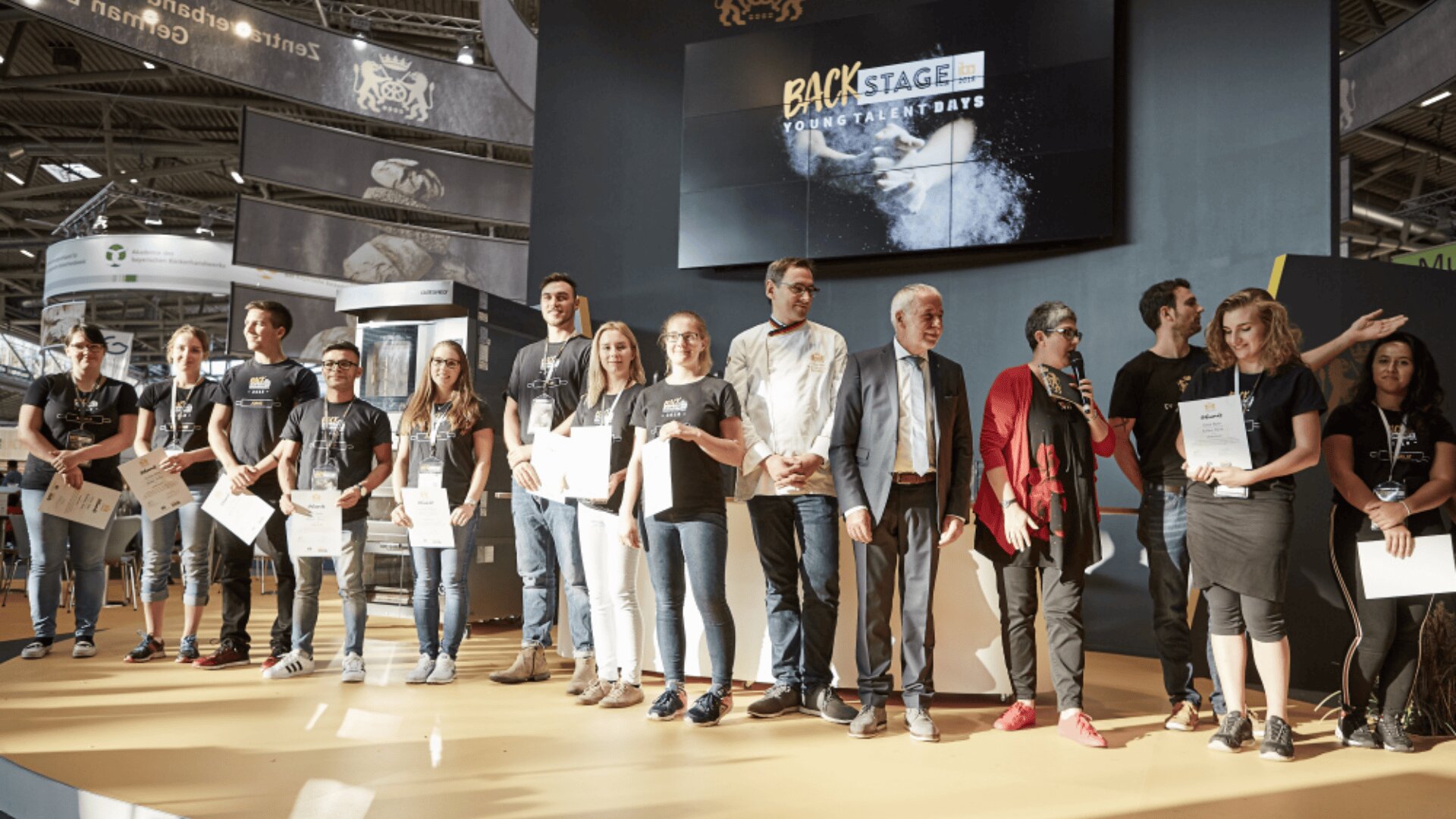 Eine Gruppe von Personen steht auf einer Bühne und hält Urkunden in der Hand. In der Mitte stehen einige Redner, hinter denen auf einer Leinwand ein Banner "Backstage Young Talents Days" zu sehen ist. Die Veranstaltung scheint eine Preisverleihungs- oder Anerkennungszeremonie zu sein.